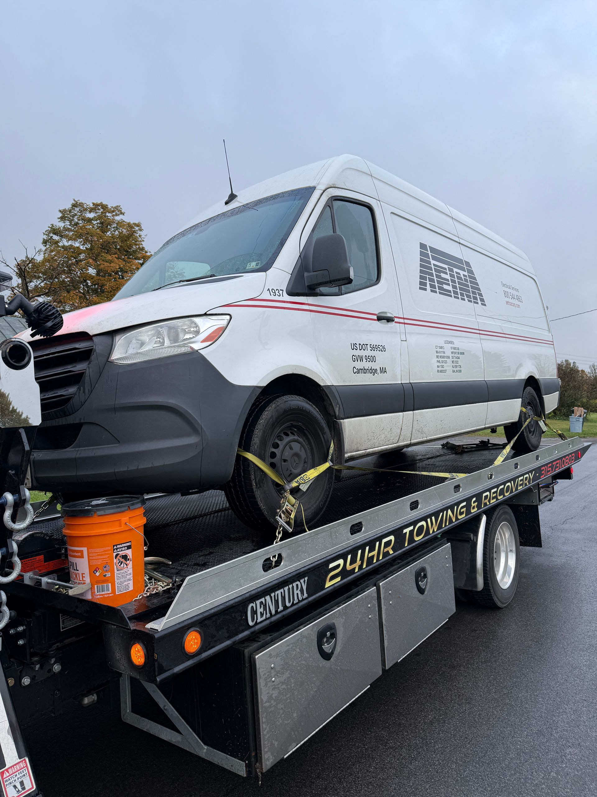 A white Mercedes-Benz cargo van secured by yellow straps onto the flatbed of a tow truck on an overcast day.