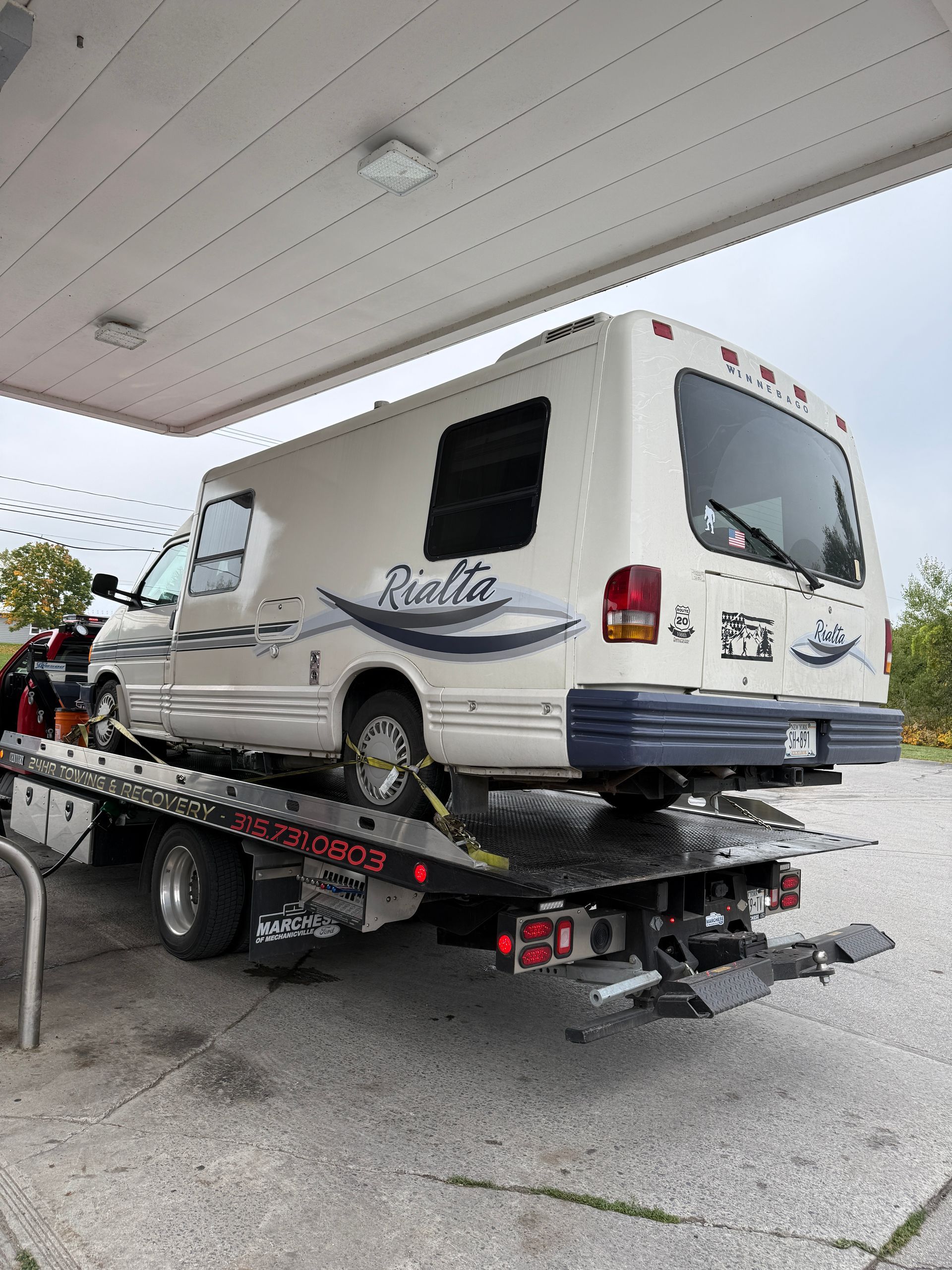 A white Rialta camper van secured to the back of a flatbed tow truck at a gas station.