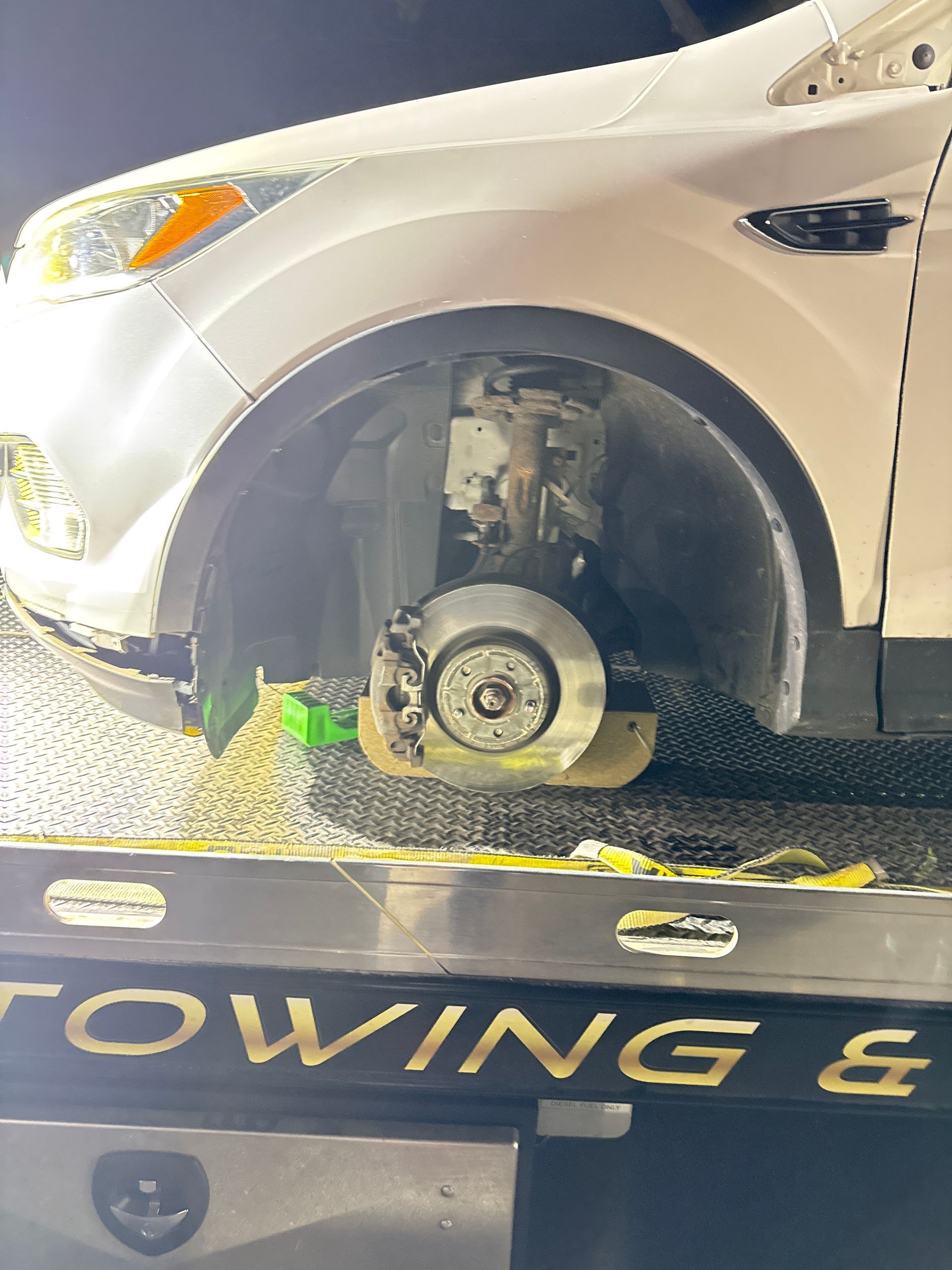 A white car without a front tire sits on the metal bed of a tow truck.