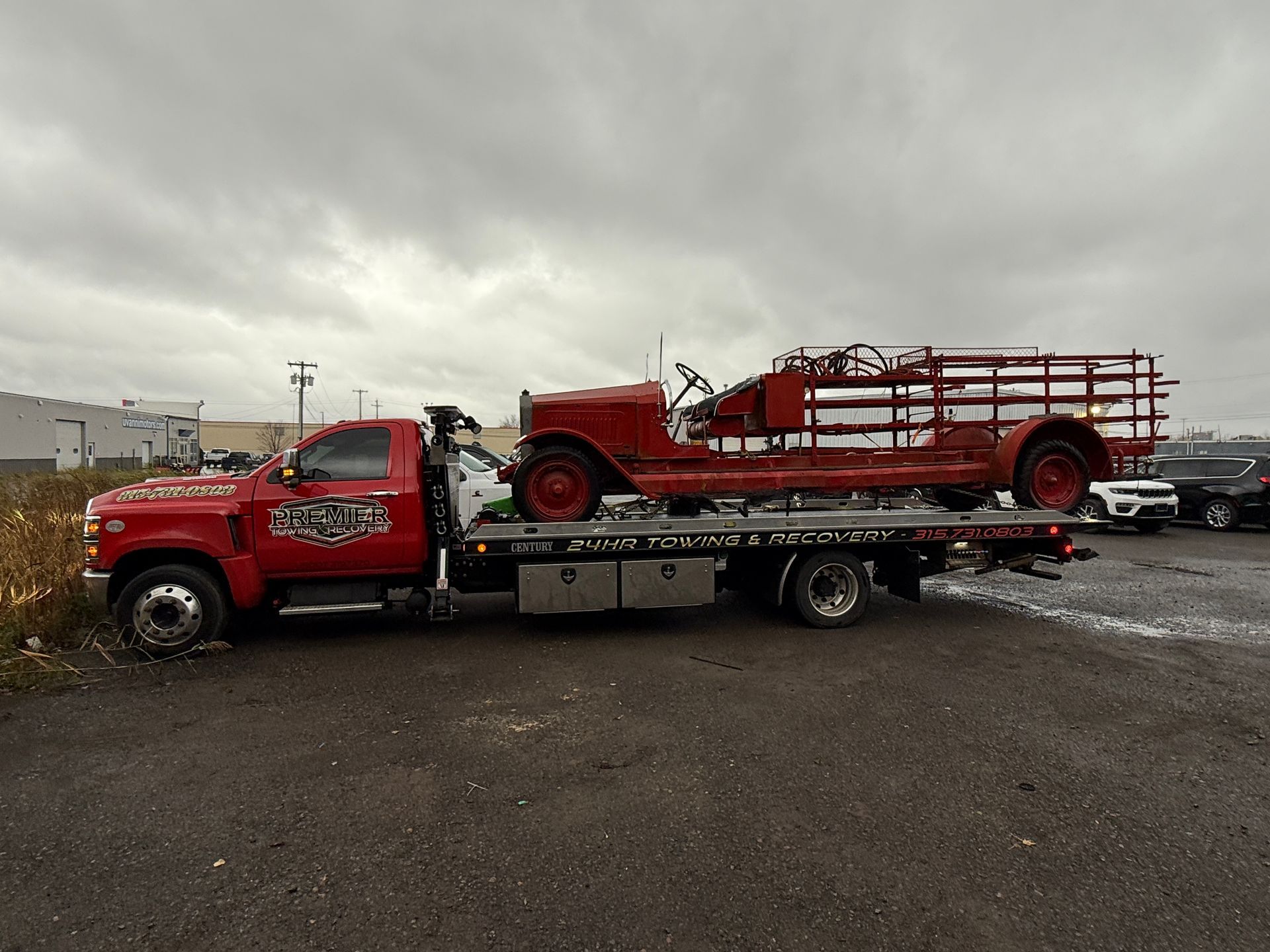 A bright red tow truck carries a vintage red fire engine on its flatbed in an overcast parking lot.