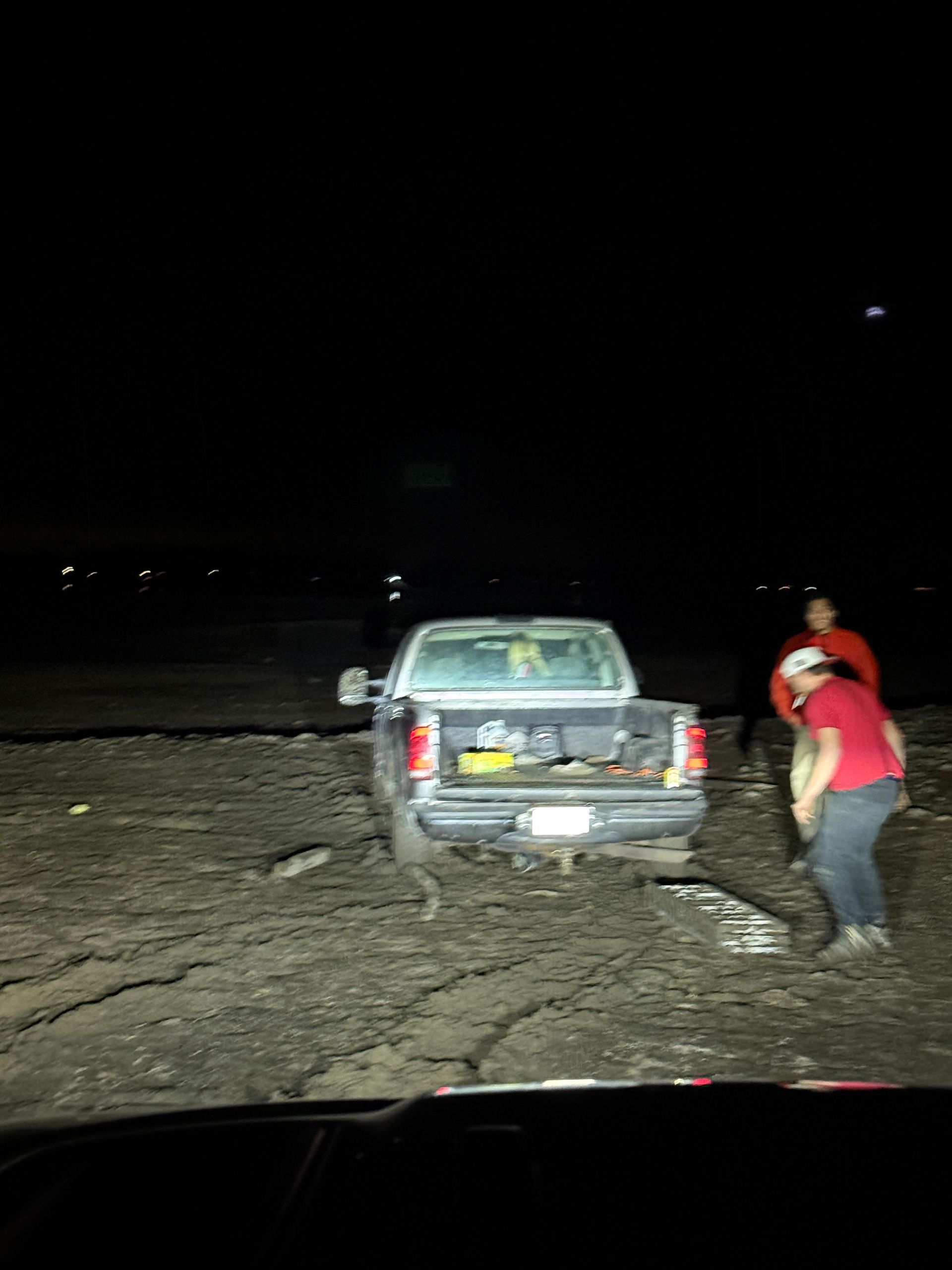 Two people in red shirts stand behind a dark pickup truck parked on a dry, cracked, and dark terrain at night.