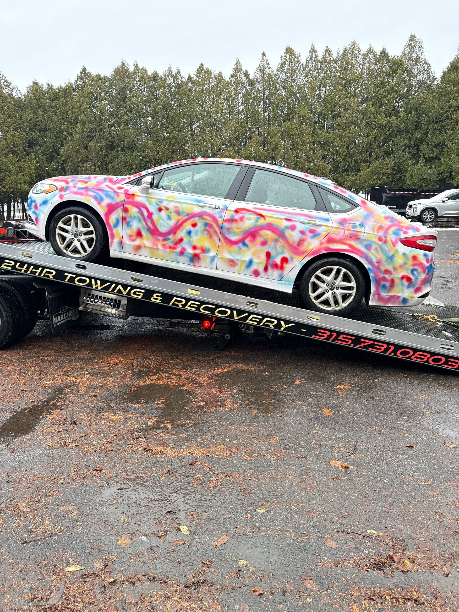 A white sedan covered in colorful, abstract graffiti paint, secured on the bed of a tow truck in an outdoor lot.