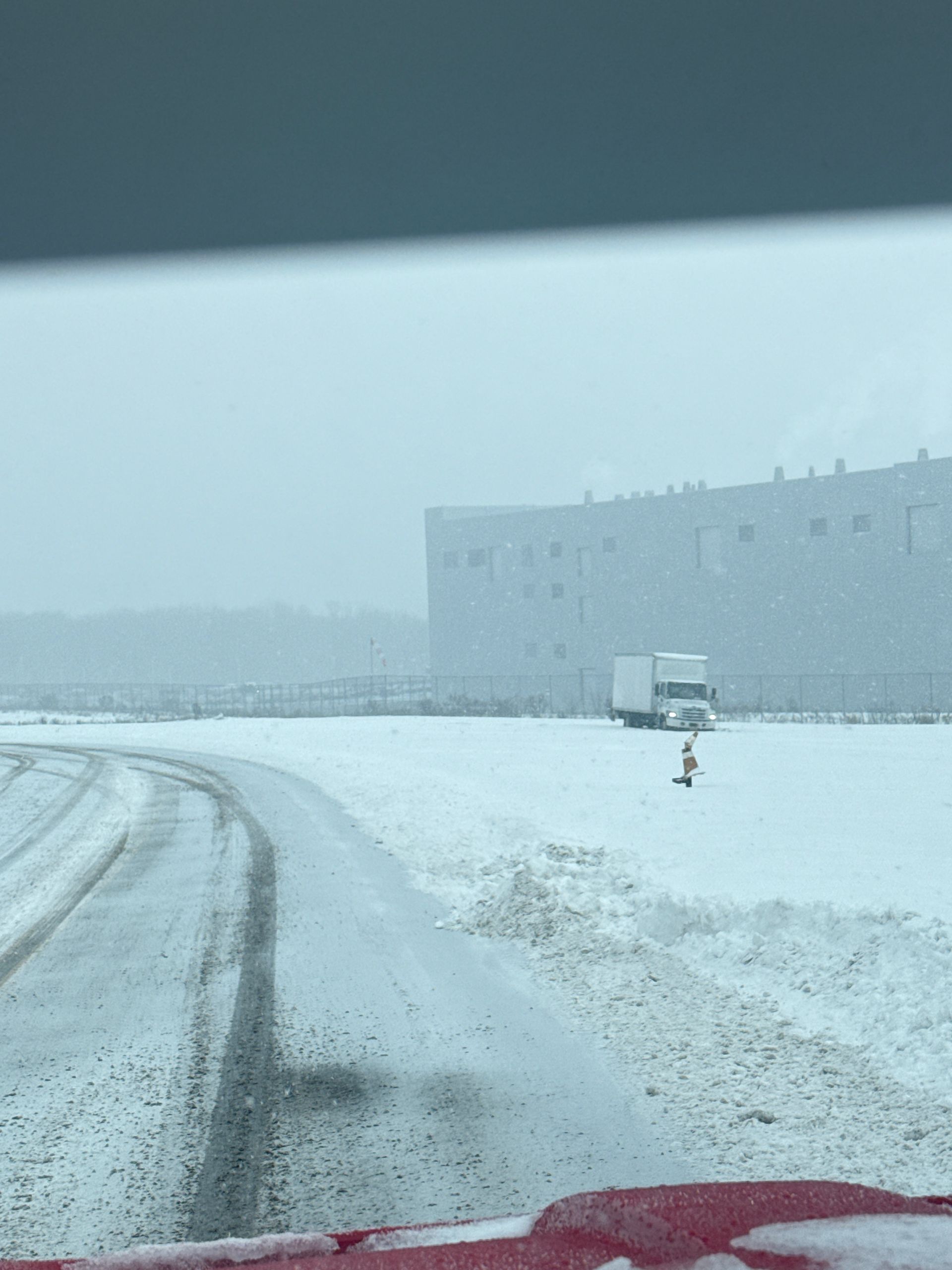 A white semi-truck is parked in a vast, snow-covered field next to a large industrial building on a snowy, overcast day.