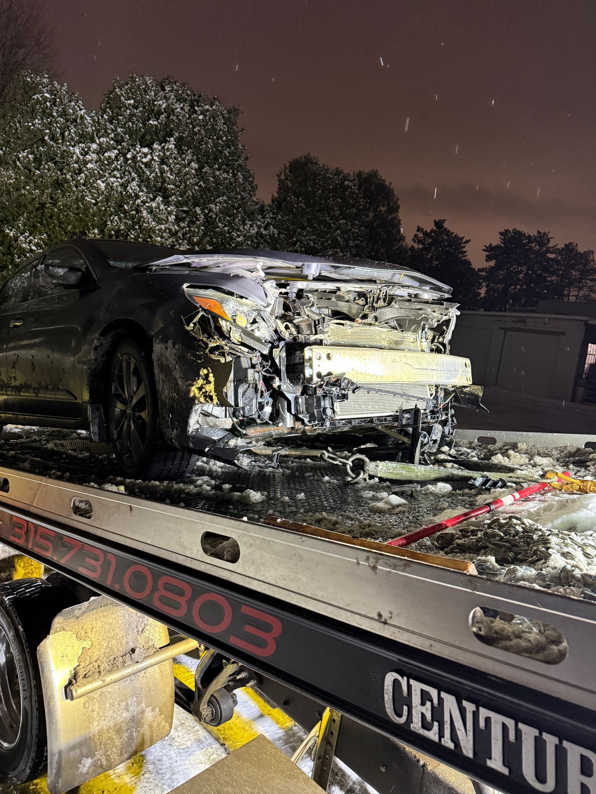 A heavily damaged car with a crushed front end secured on the back of a Century tow truck at night.