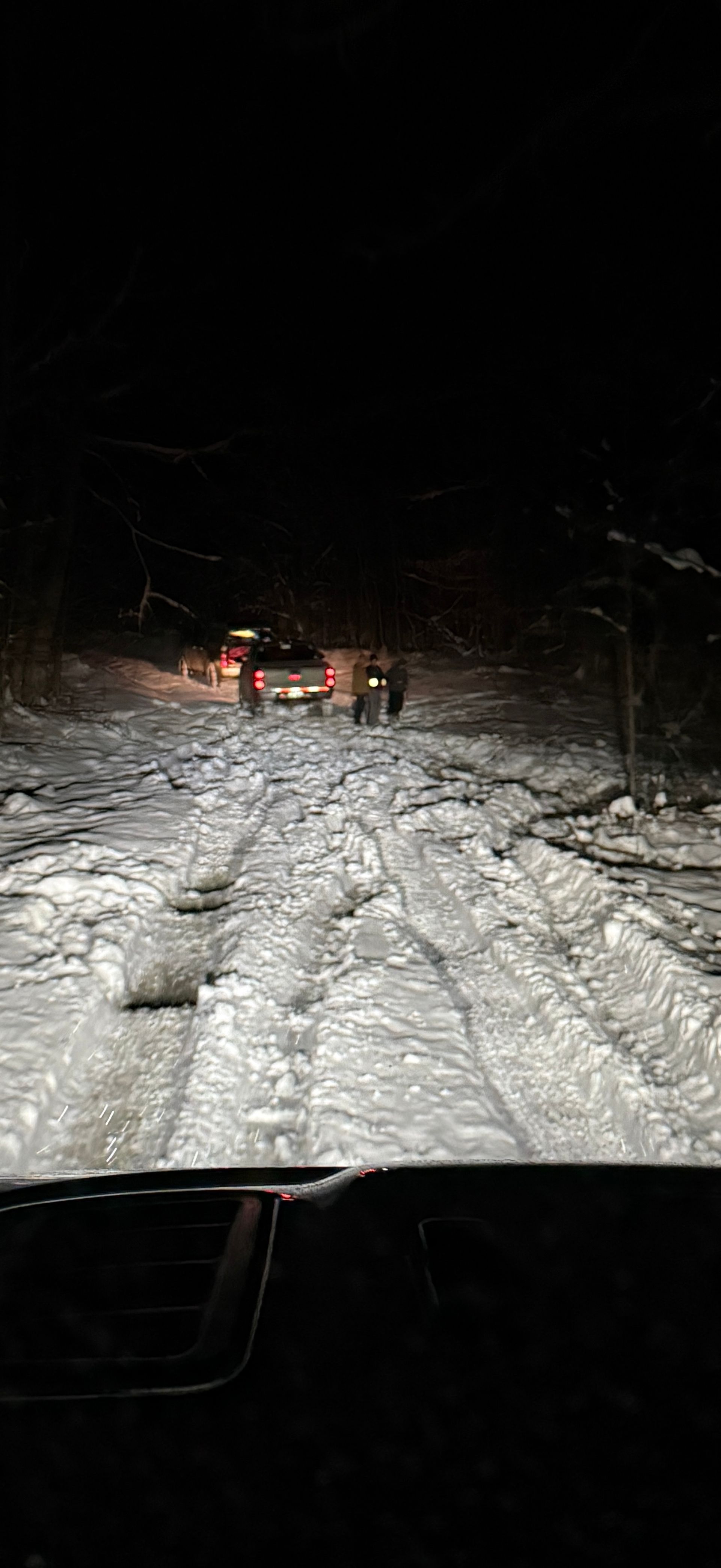 A vehicle drives along a snowy, unpaved road at night toward another car with its taillights visible in the distance.