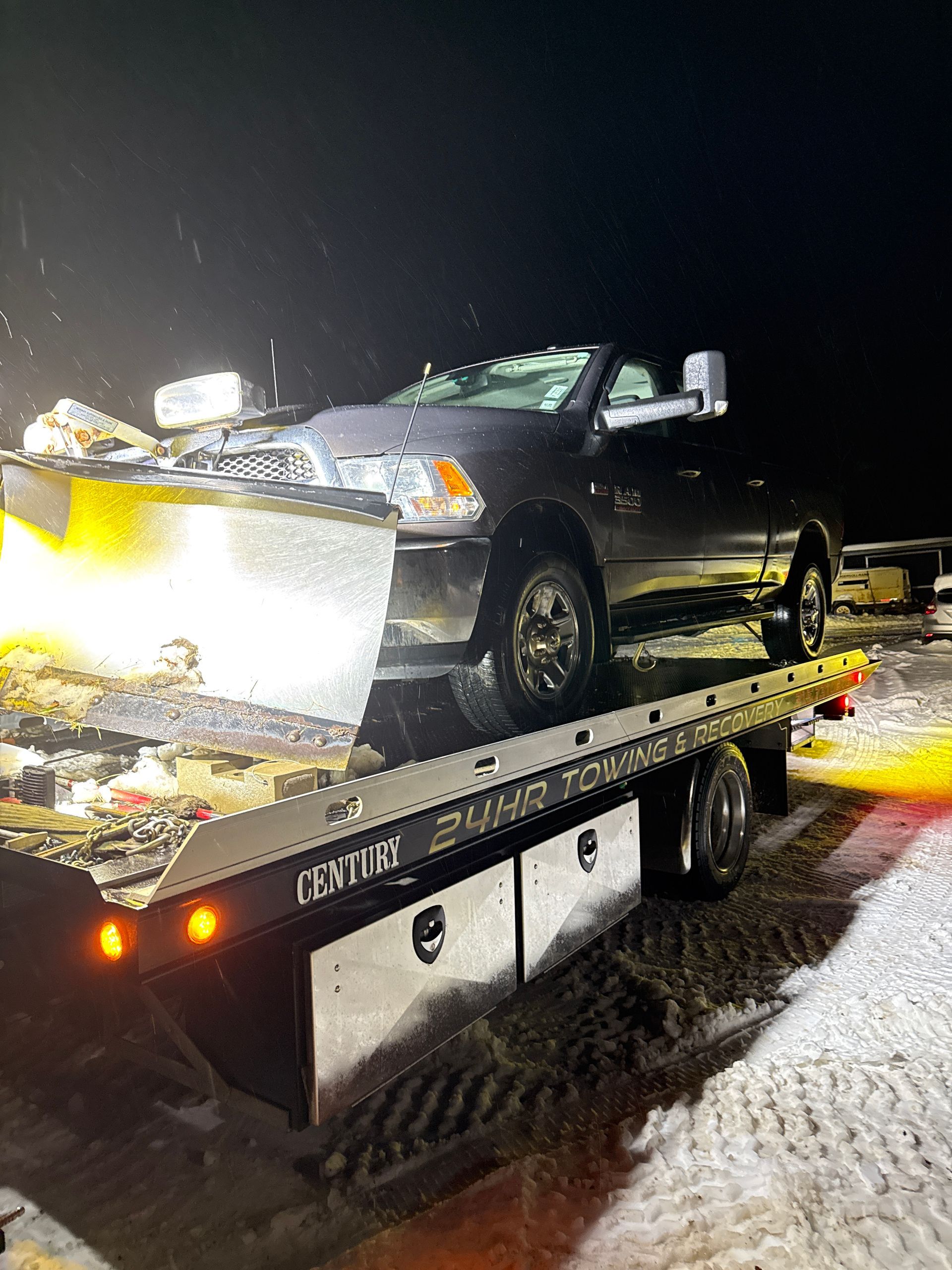 A dark pickup truck with a large snowplow blade attached is loaded onto the flatbed of a tow truck at night.