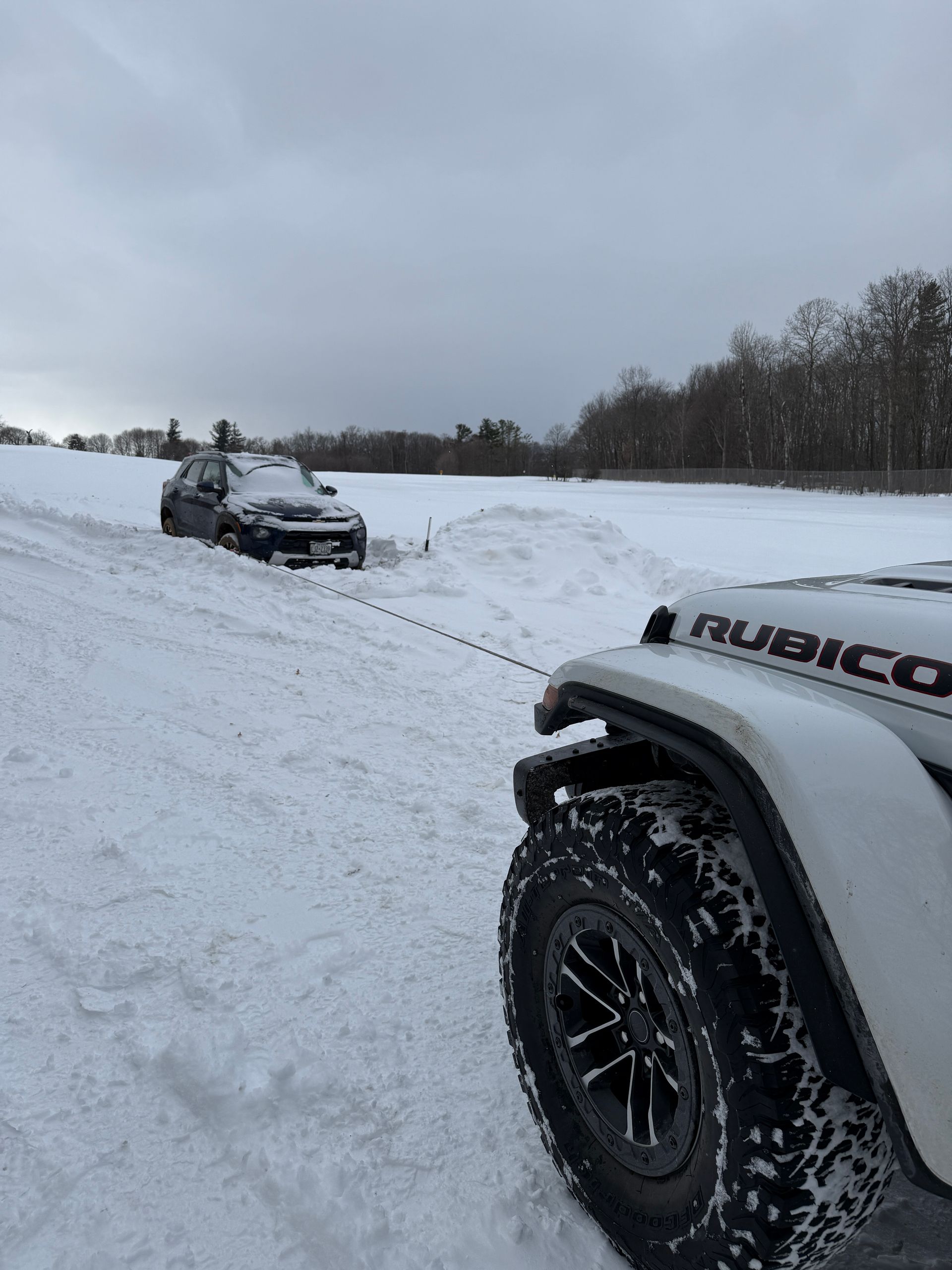 A white Jeep Rubicon pulling a stuck car out of deep snow in a field.
