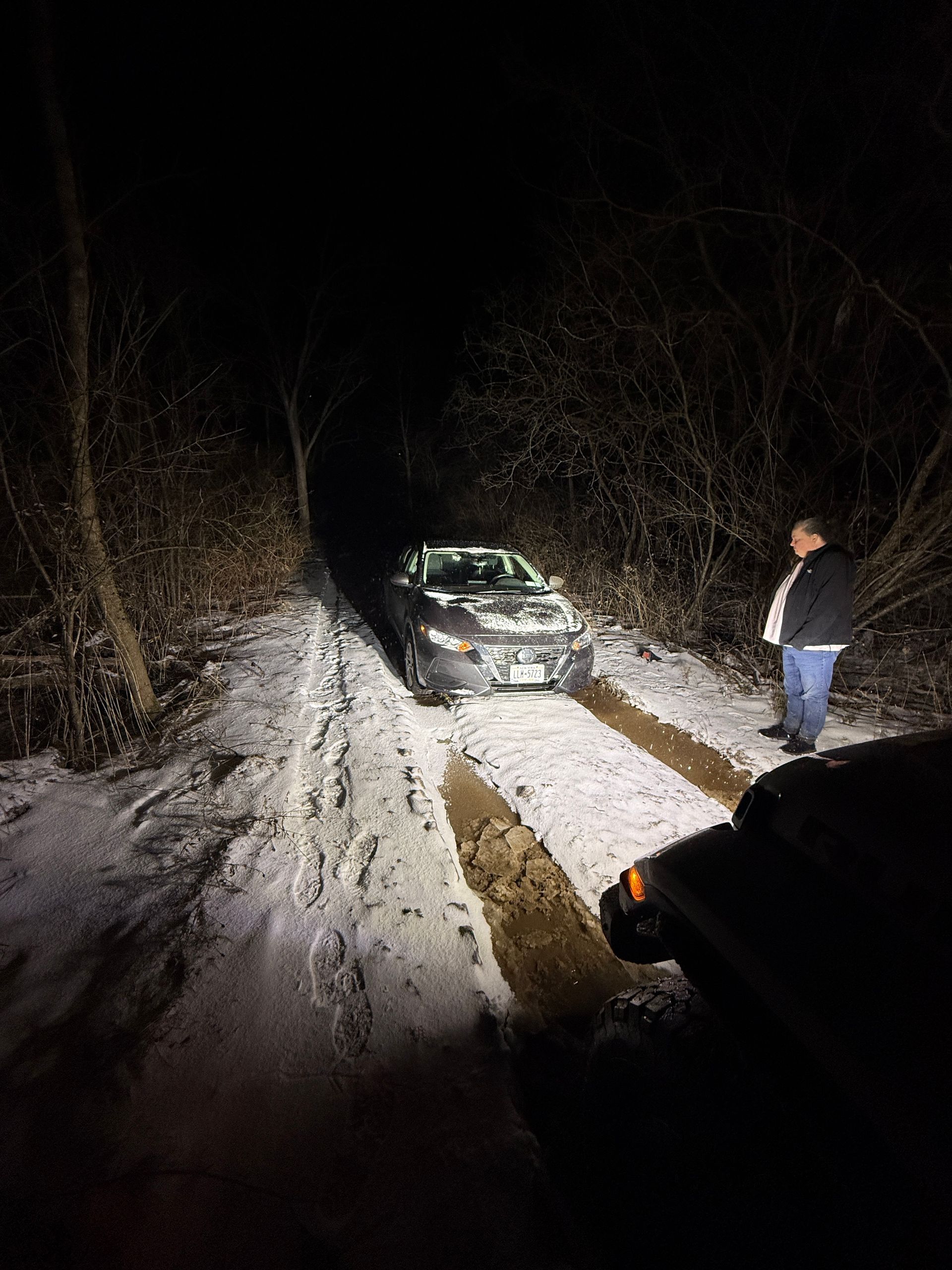 A car stuck in deep snow on a dark, wooded trail at night, with a person standing nearby in the headlights.