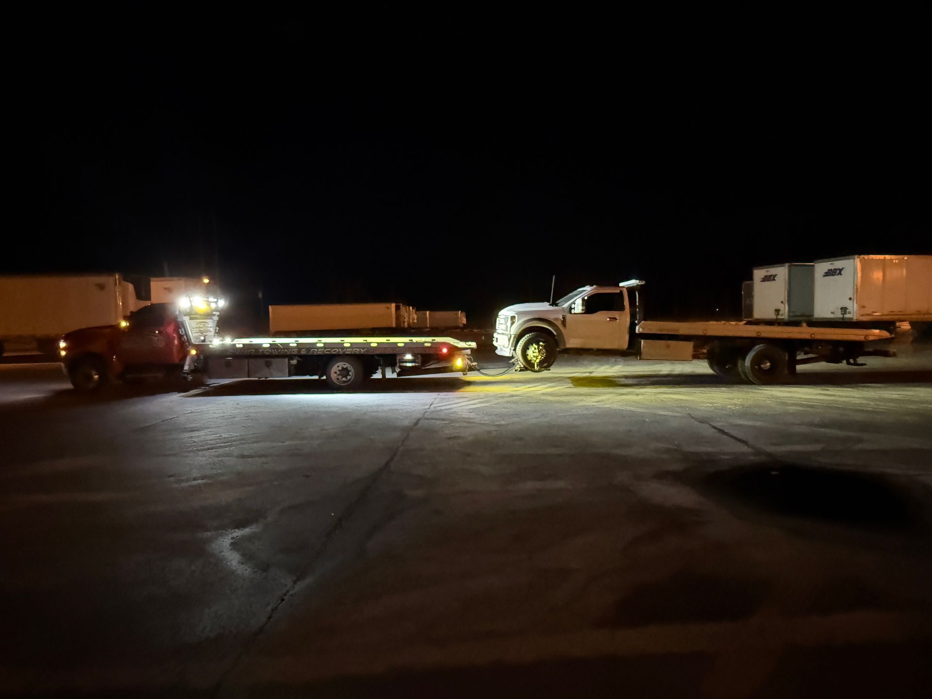 A flatbed tow truck pulls a white pickup truck across a dark, paved lot at night.