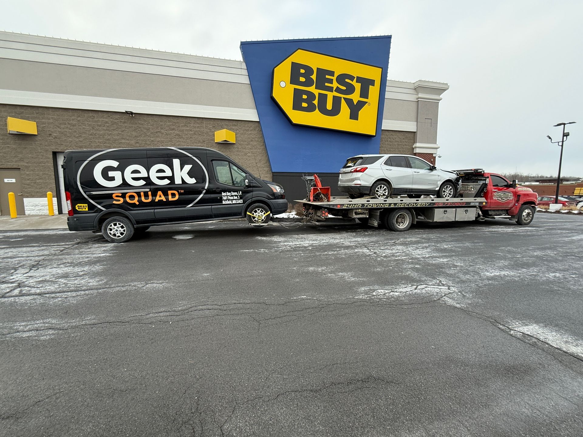 A Geek Squad van towing a flatbed truck with a silver sedan on it in front of a Best Buy store.