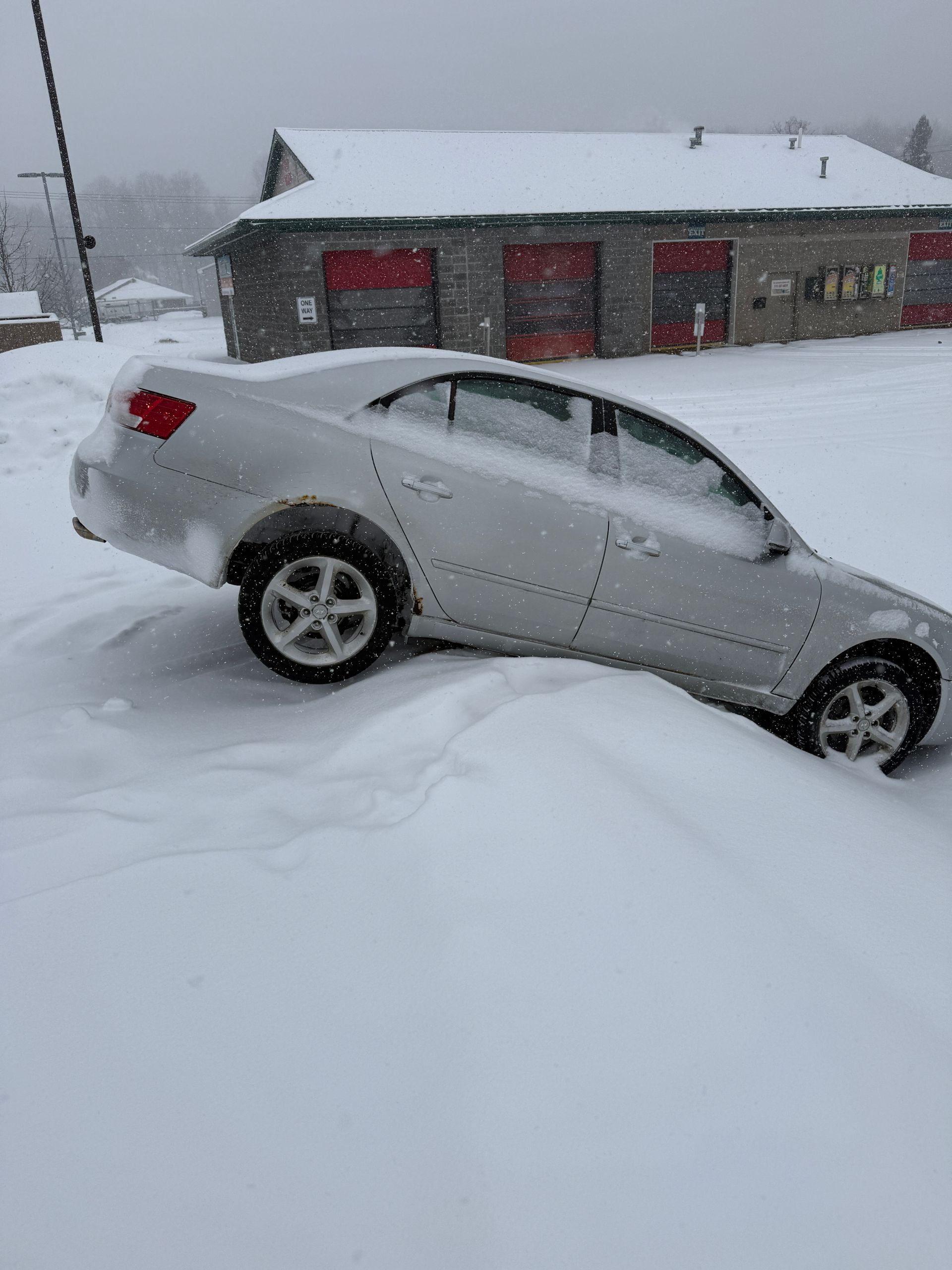 A silver sedan stuck in a deep snowbank in front of a building with red garage doors during a snowfall.