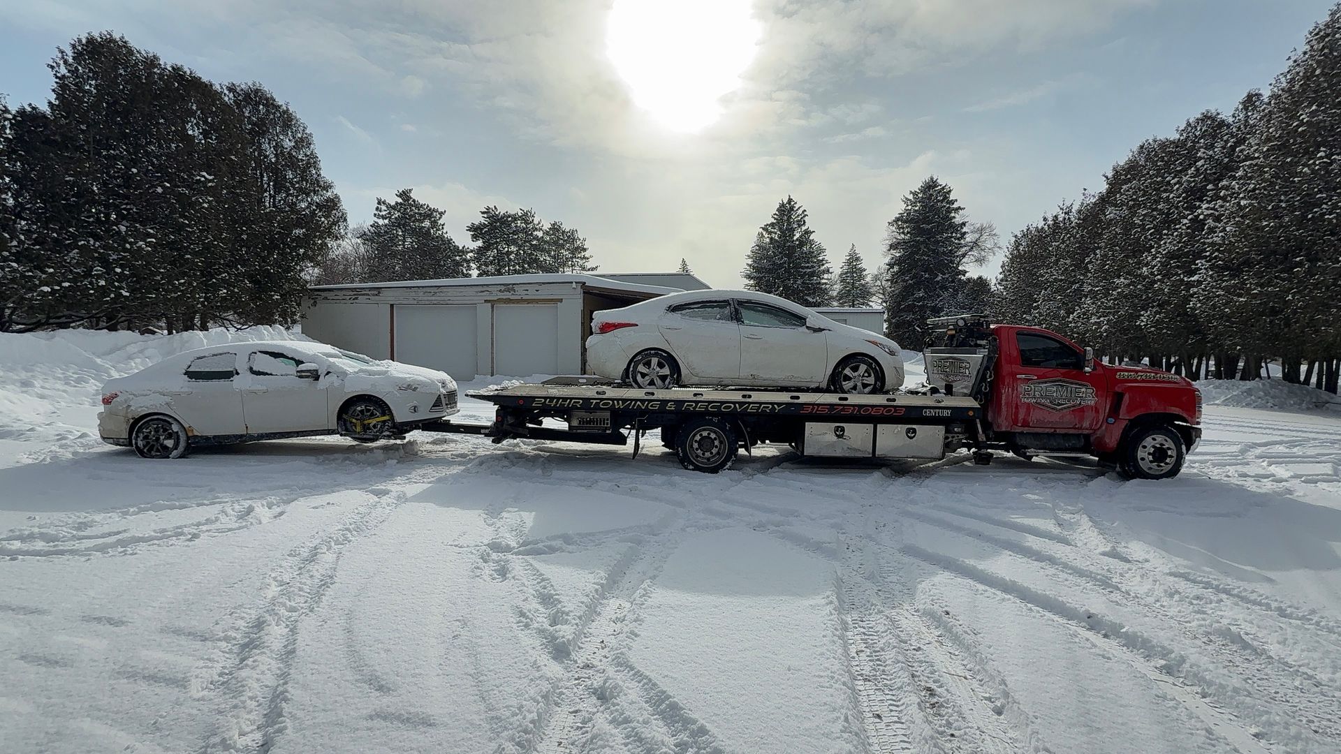 A red tow truck carries a white sedan while towing a second white sedan through a snowy, wooded area on a sunny day.