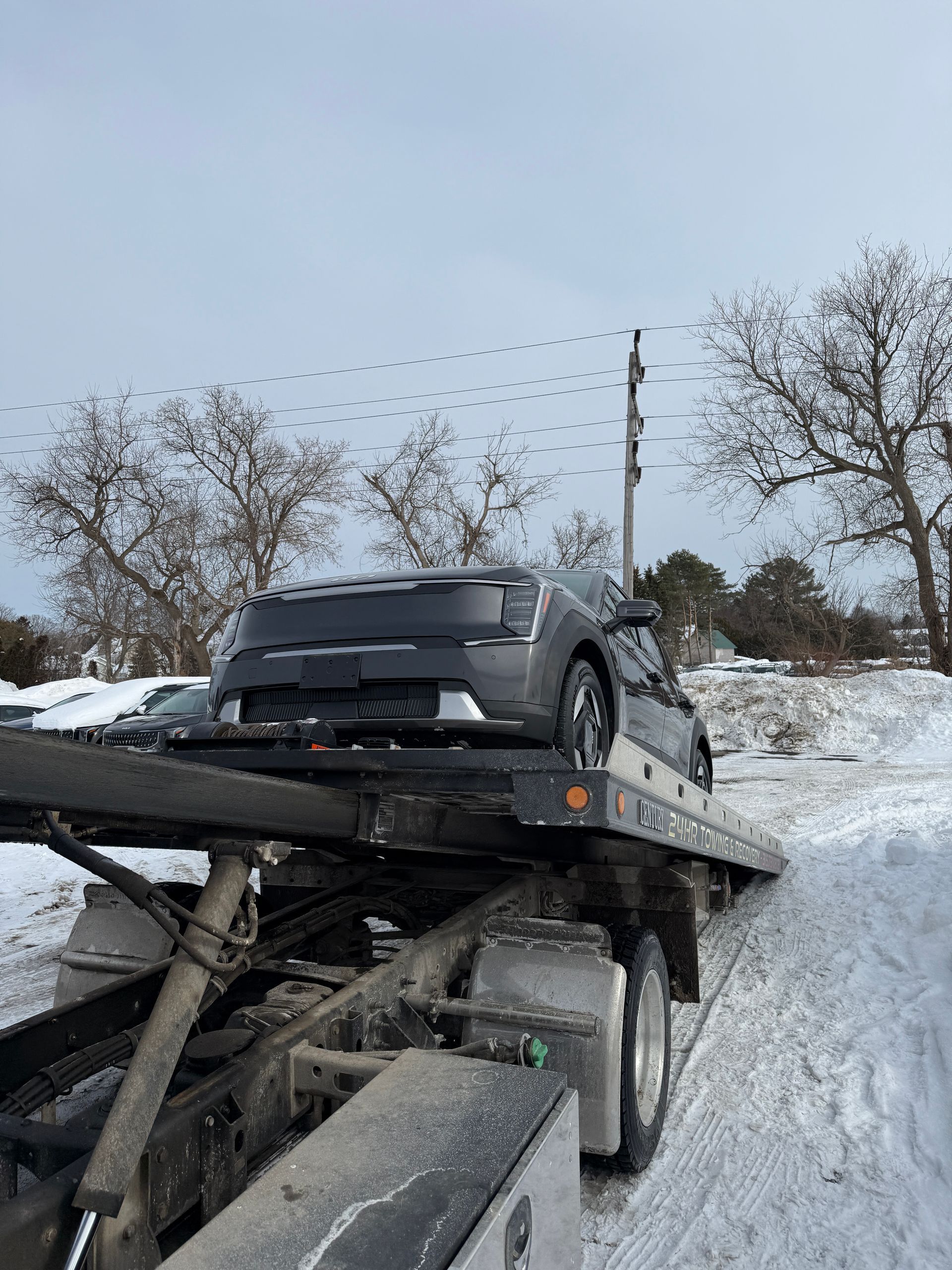 A gray Kia EV9 vehicle being transported on a flatbed tow truck on a snow-covered road.