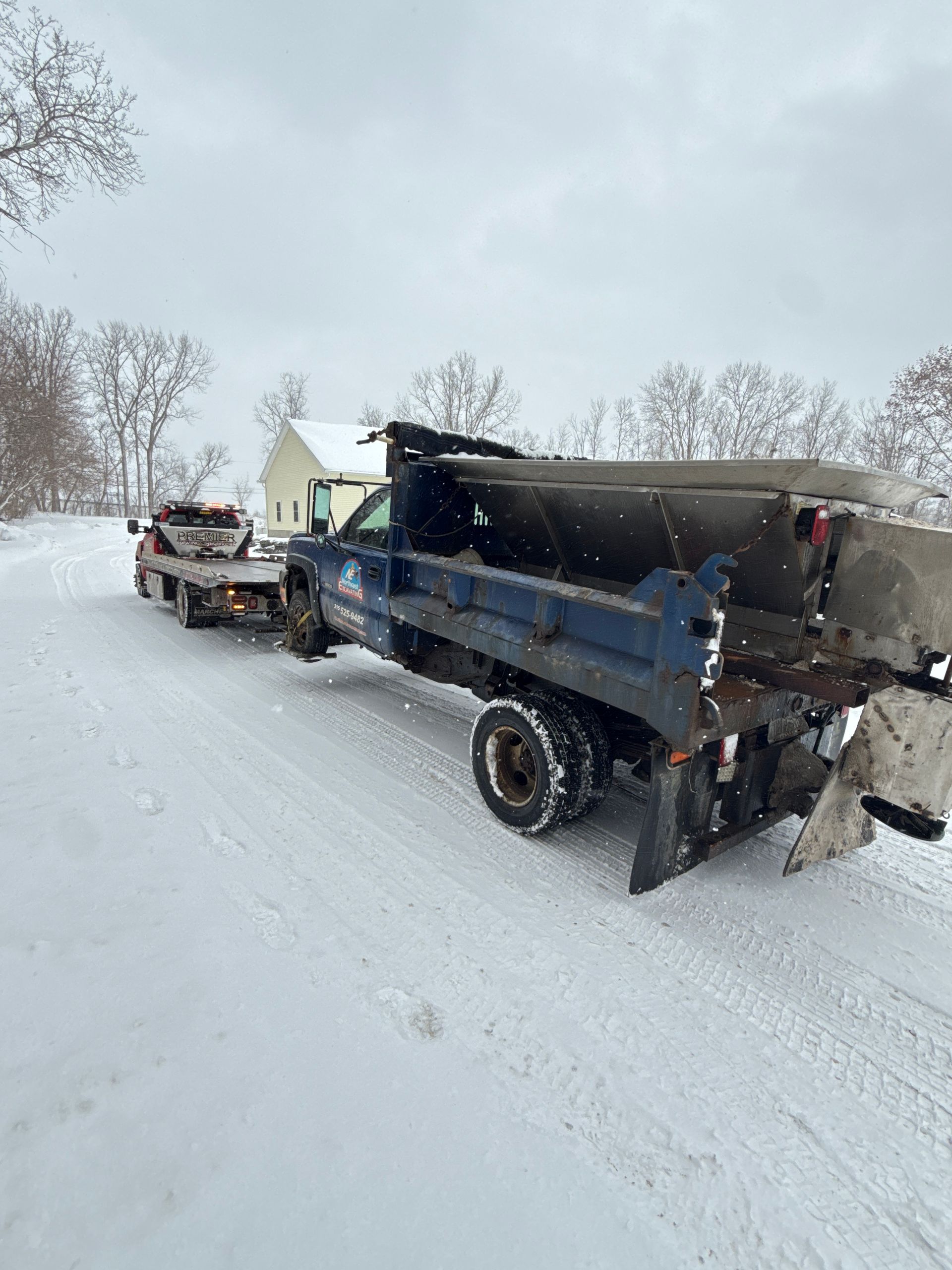 A blue dump truck with a salt spreader pulling a flatbed trailer on a snow-covered road.