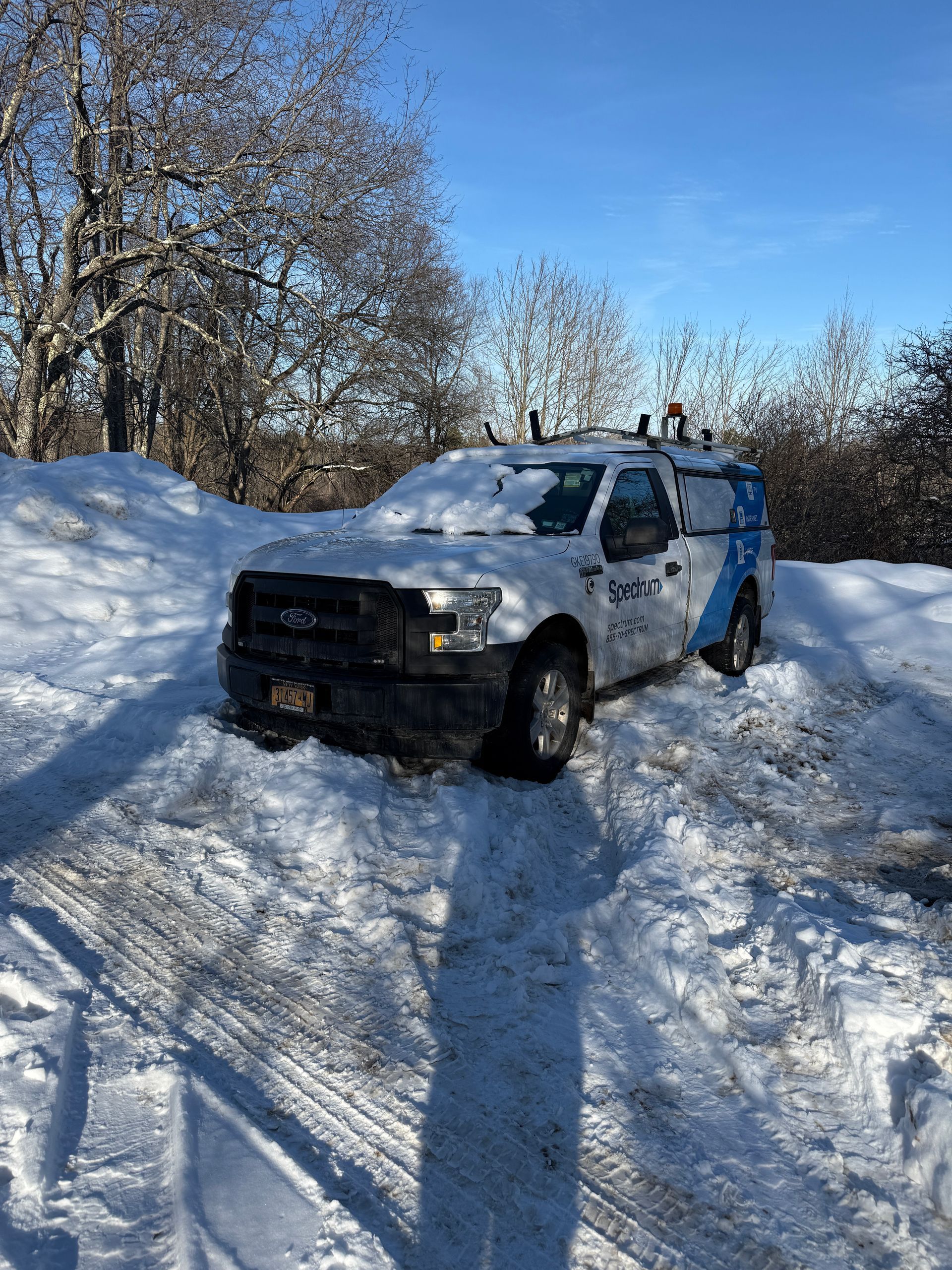A white service truck covered in snow parked on a snowy, sunny landscape with trees in the background.