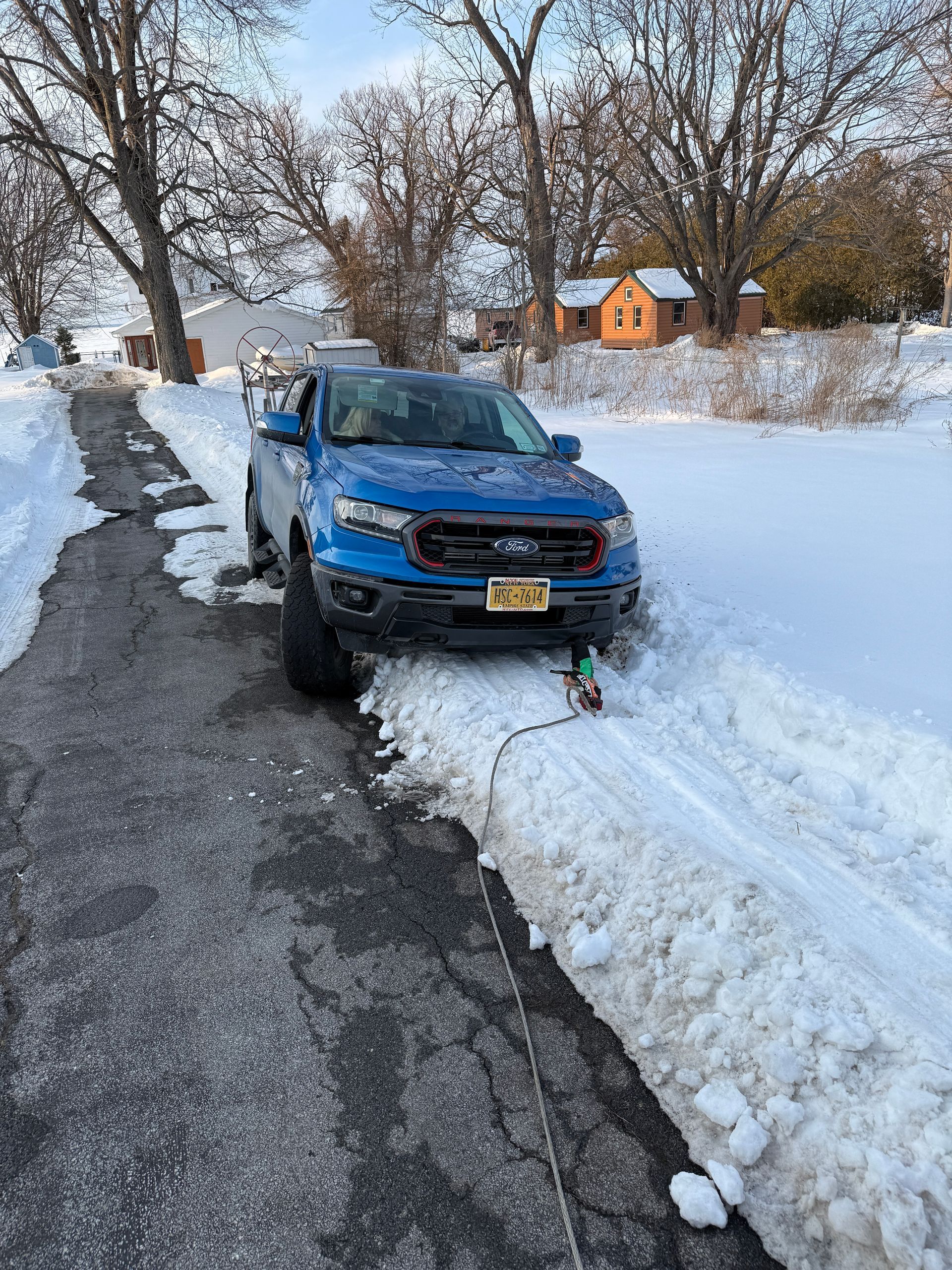 A blue Ford Ranger pickup truck stuck in deep snow on the side of a partially cleared paved driveway.