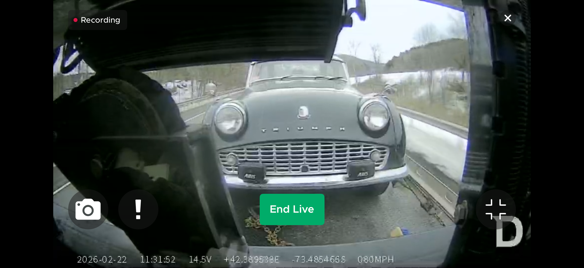 A dashcam view of a vintage dark green Triumph sports car being transported on the back of a flatbed truck.