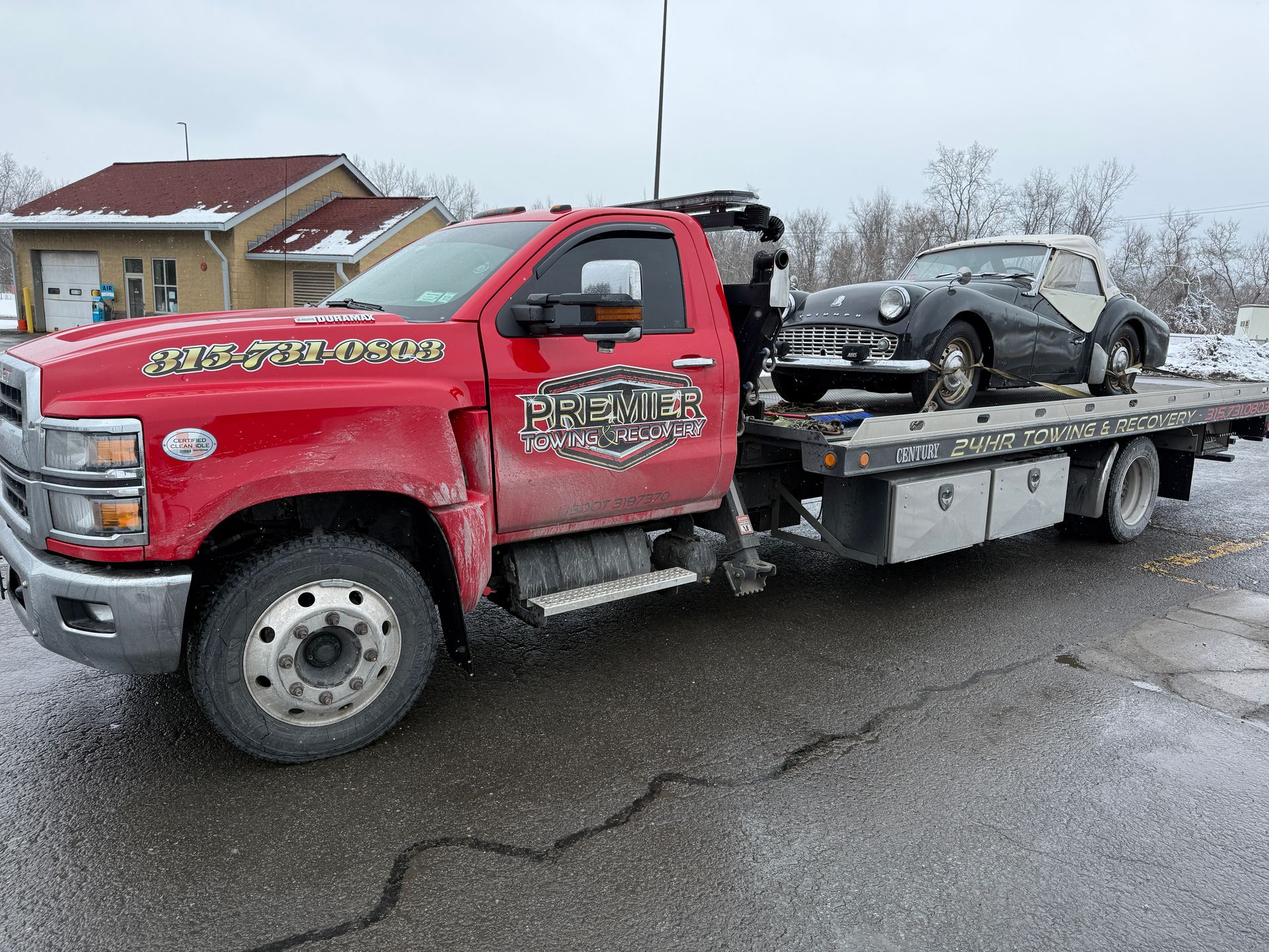 A red flatbed tow truck carrying a black vintage convertible on its back in a snowy, outdoor setting.