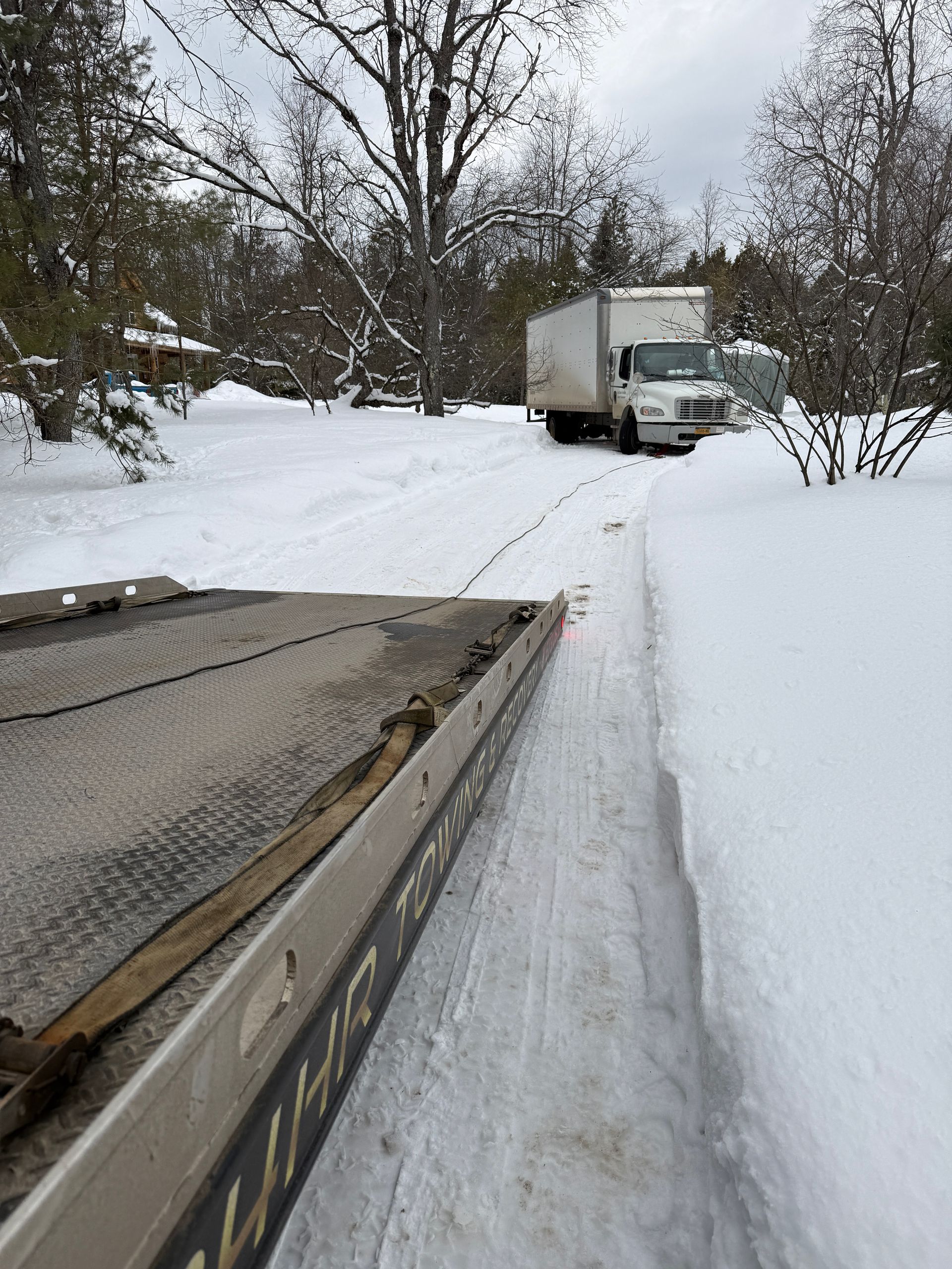 A white box truck parked on a snow-covered path, viewed from the bed of a trailer in the foreground.