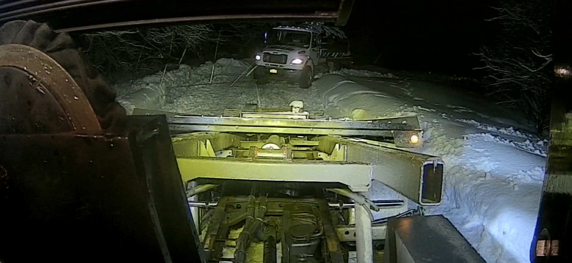 View from inside a cab showing a snow plow clearing a snowy, dark road at night with another vehicle approaching ahead.