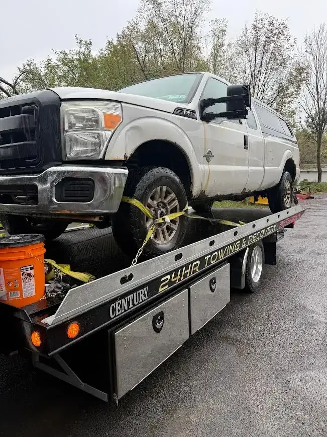 A white Ford Super Duty truck is secured with yellow straps to the flatbed of a 24-hour tow truck on an overcast day.
