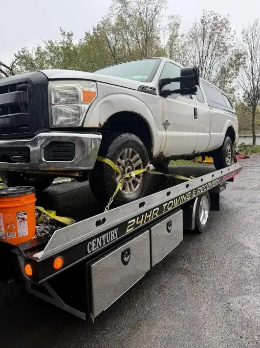 A white pickup truck secured with yellow tie-down straps on a flatbed tow truck parked outdoors.