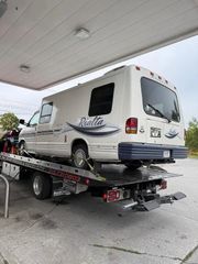 A white Rialta motorhome secured on a flatbed tow truck at a gas station.