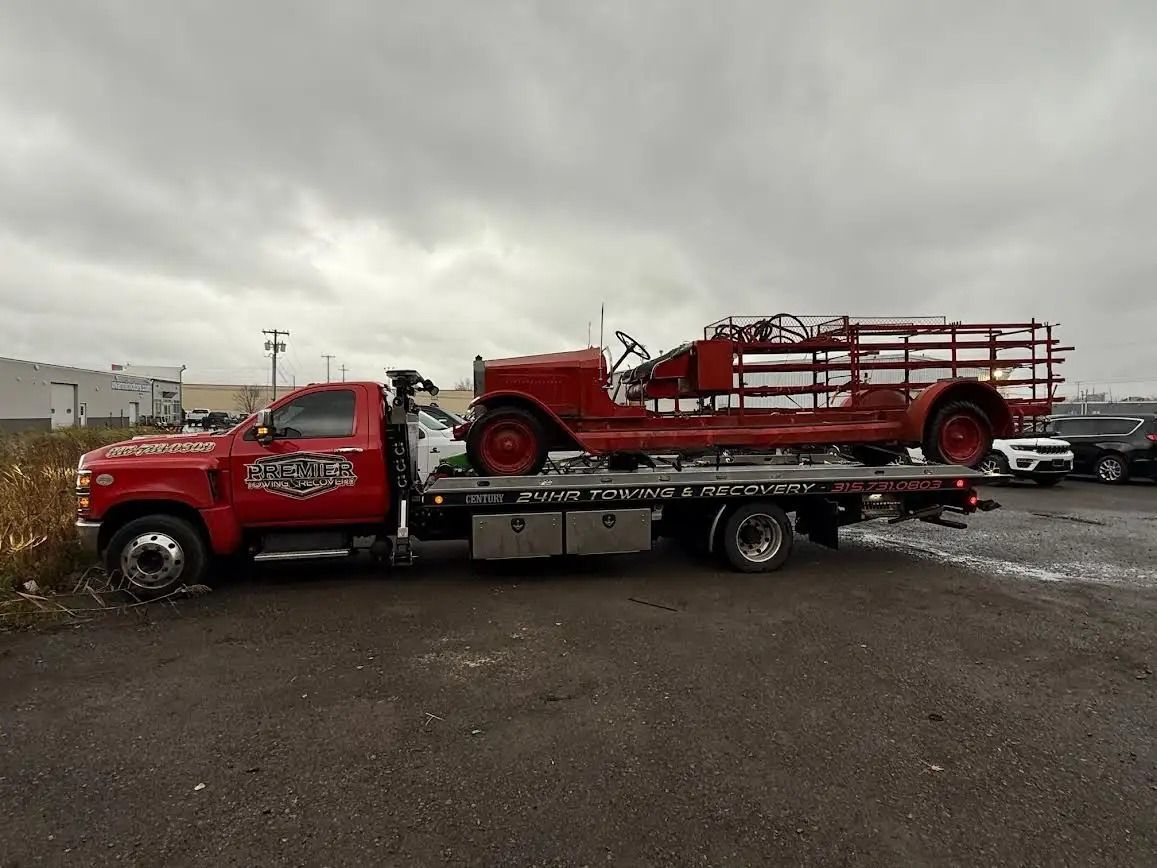 A red flatbed tow truck carries a vintage red fire engine in an outdoor parking lot under a cloudy sky.