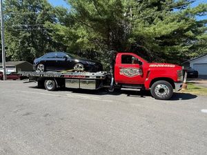 A bright red tow truck carrying a black sedan parked on a gravel lot in front of trees.