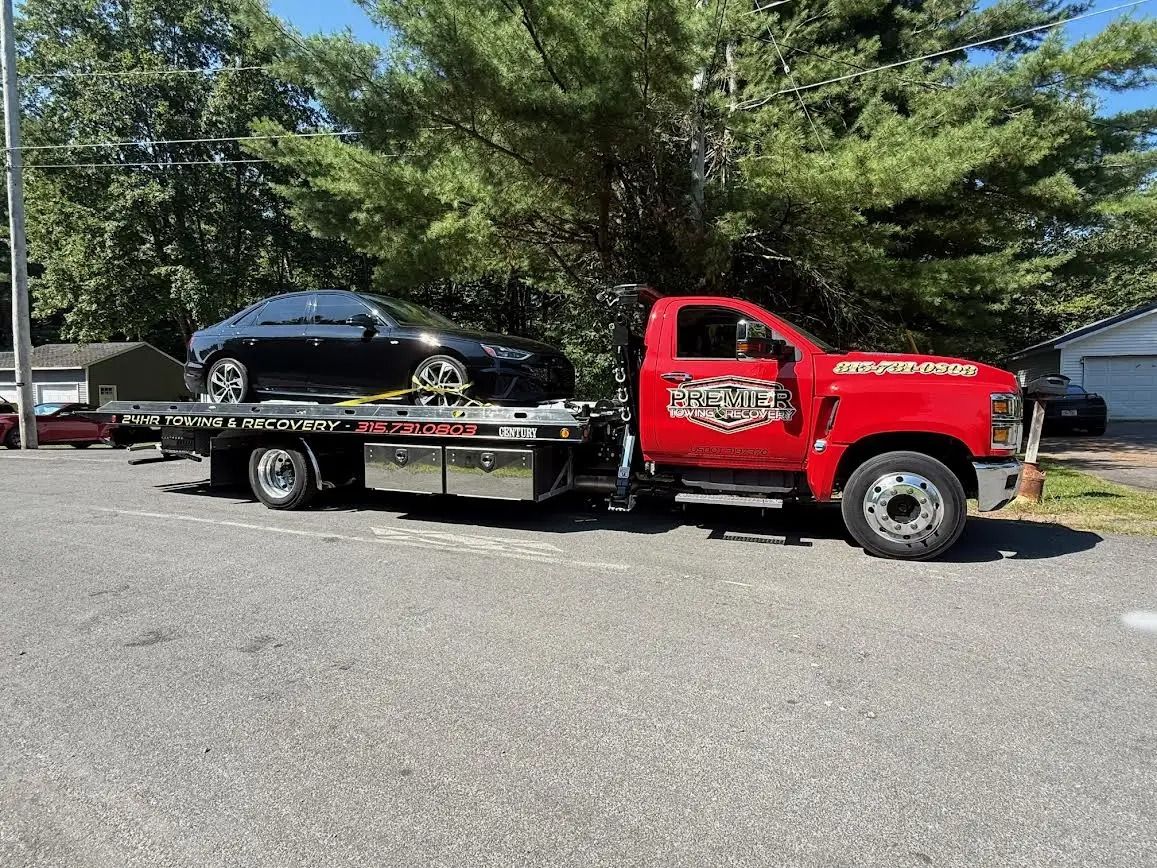 A bright red tow truck carries a black sedan on its flatbed, parked on an asphalt lot in front of trees.