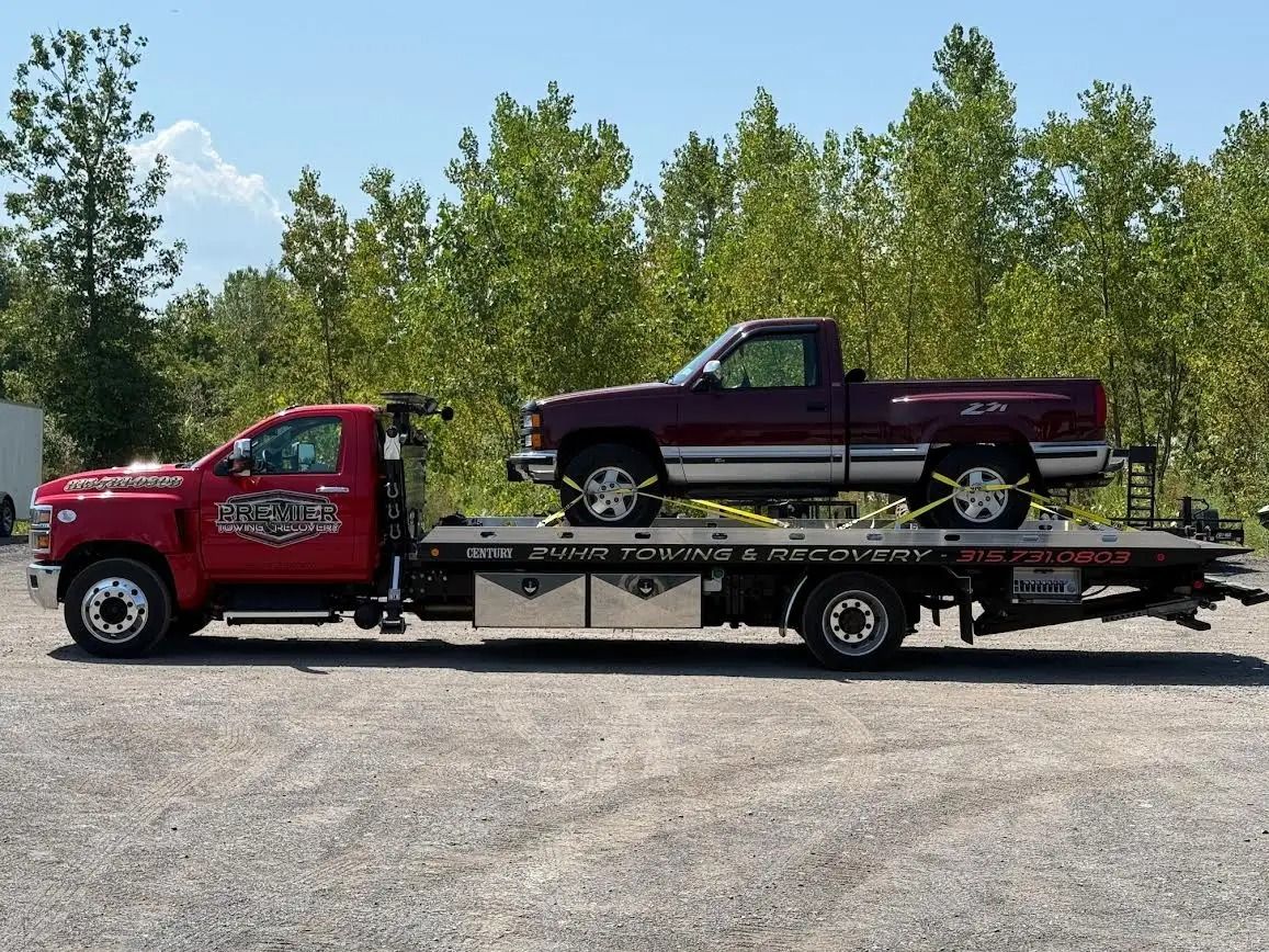 A red flatbed tow truck transporting a maroon pickup truck on a gravel lot against a backdrop of trees.