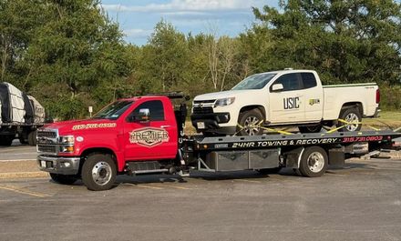 A bright red tow truck carries a white USIC pickup truck on its flatbed in a parking lot.
