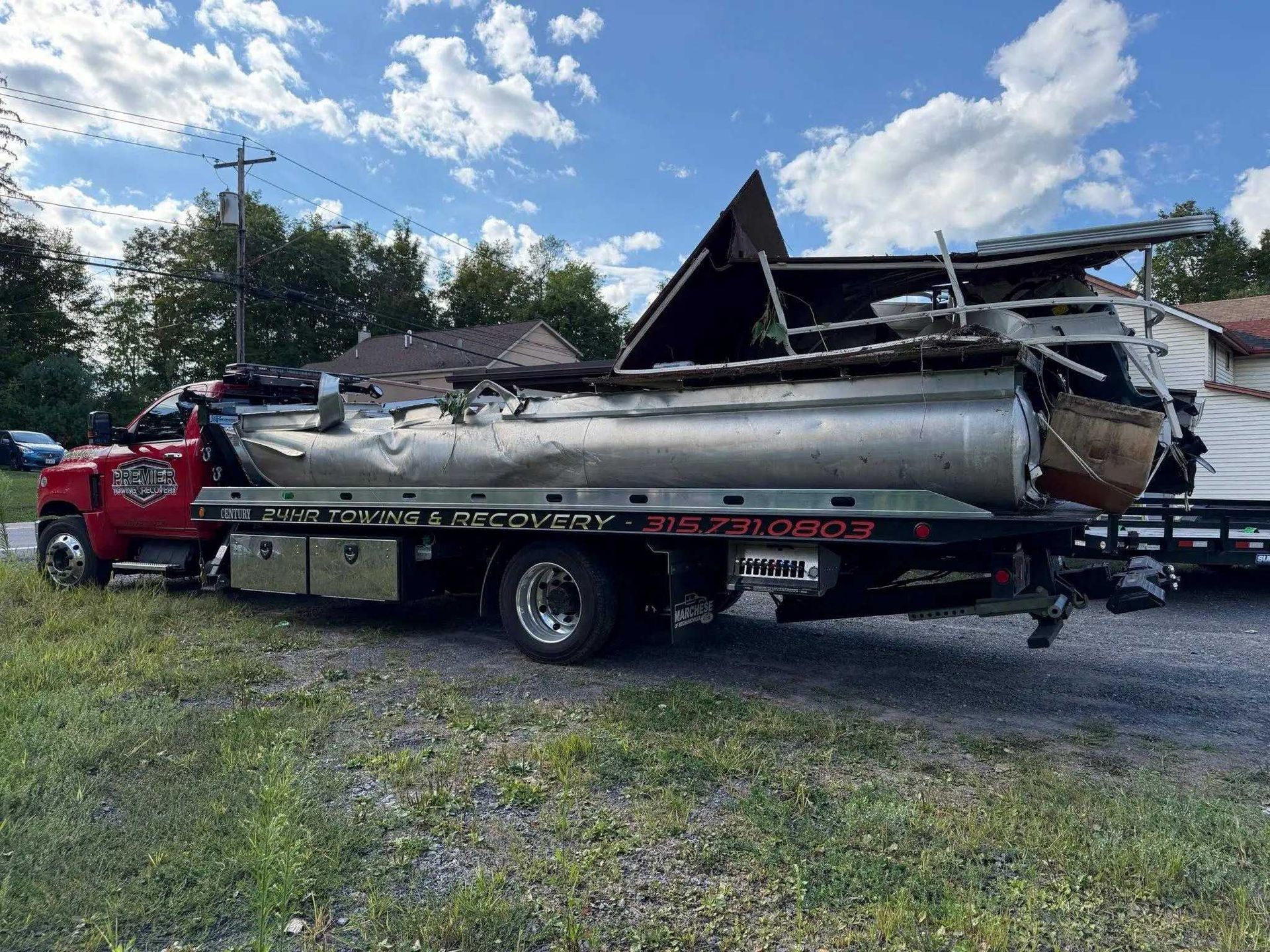 A red tow truck carries a heavily damaged, silver pontoon boat on its flatbed in a gravel lot under a blue sky.