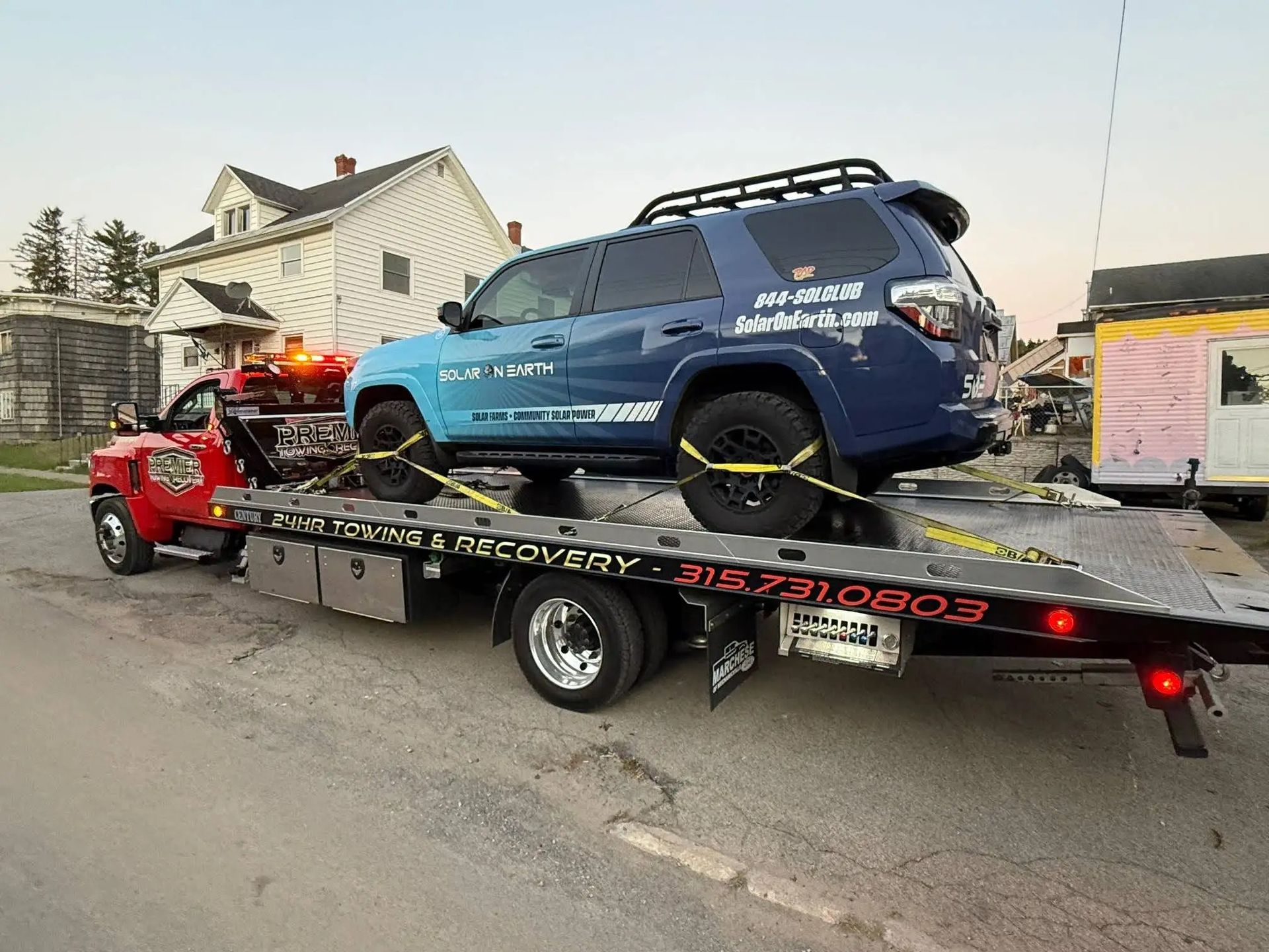 A blue SUV is secured to the flatbed of a red tow truck parked on a street in front of a house.