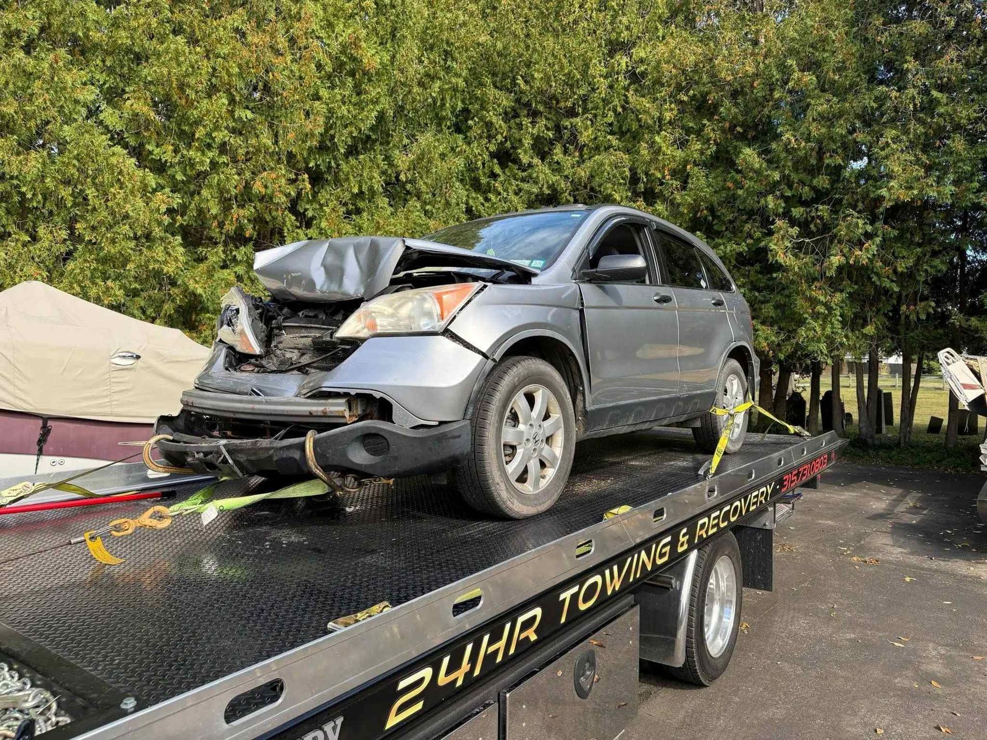 A damaged silver Honda CR-V is secured to the back of a flatbed tow truck parked in front of trees.