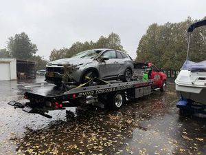 A silver SUV is strapped to the flatbed of a tow truck in a rainy, leaf-covered lot with a boat nearby.