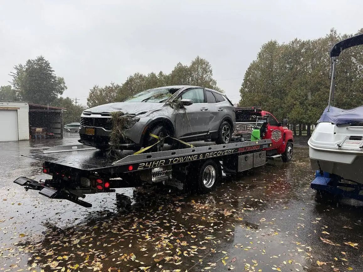 A silver SUV is loaded onto a flatbed tow truck in a rainy, leaf-covered lot with a boat nearby.