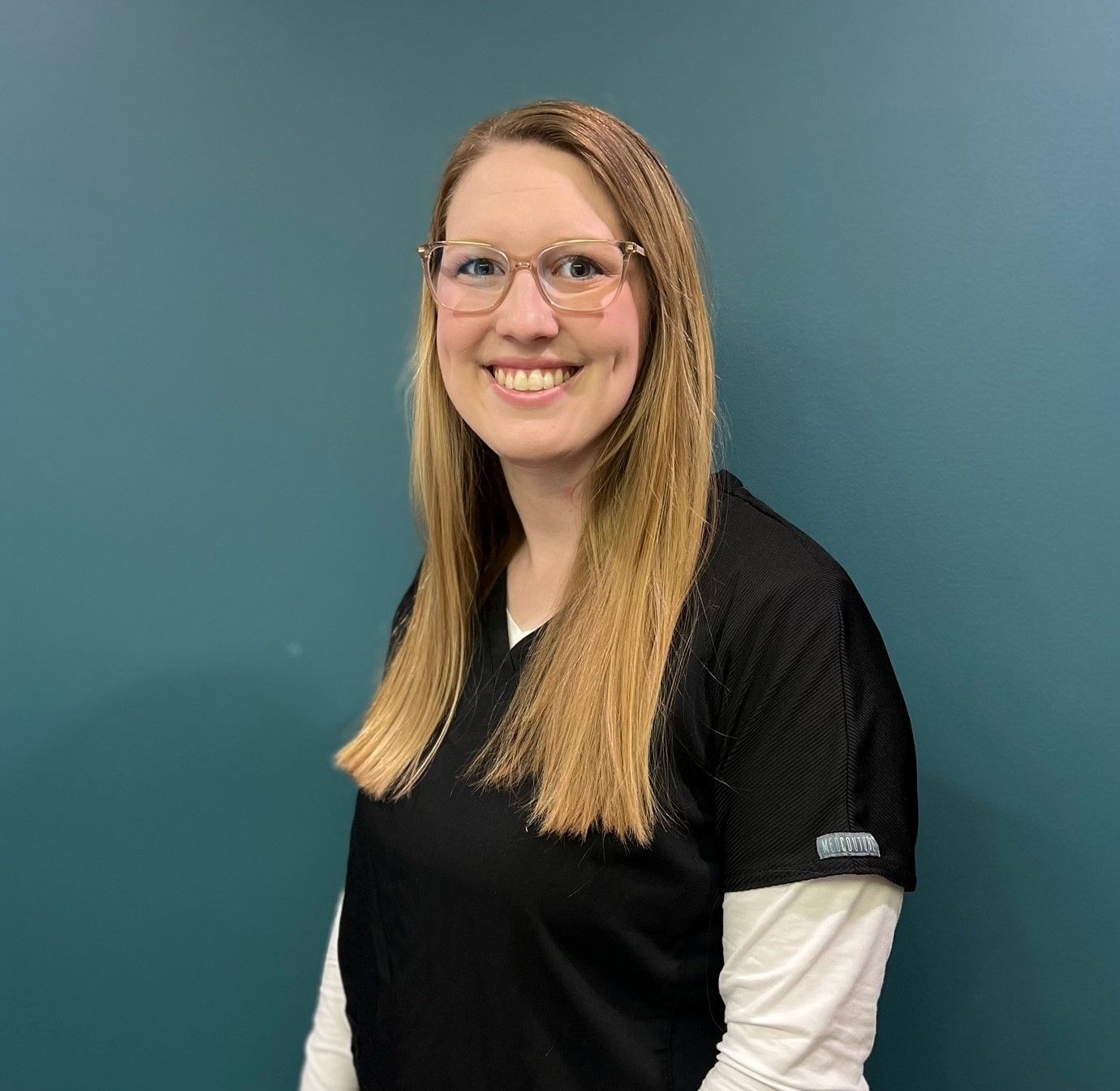 Smiling person in glasses and black shirt against a teal wall