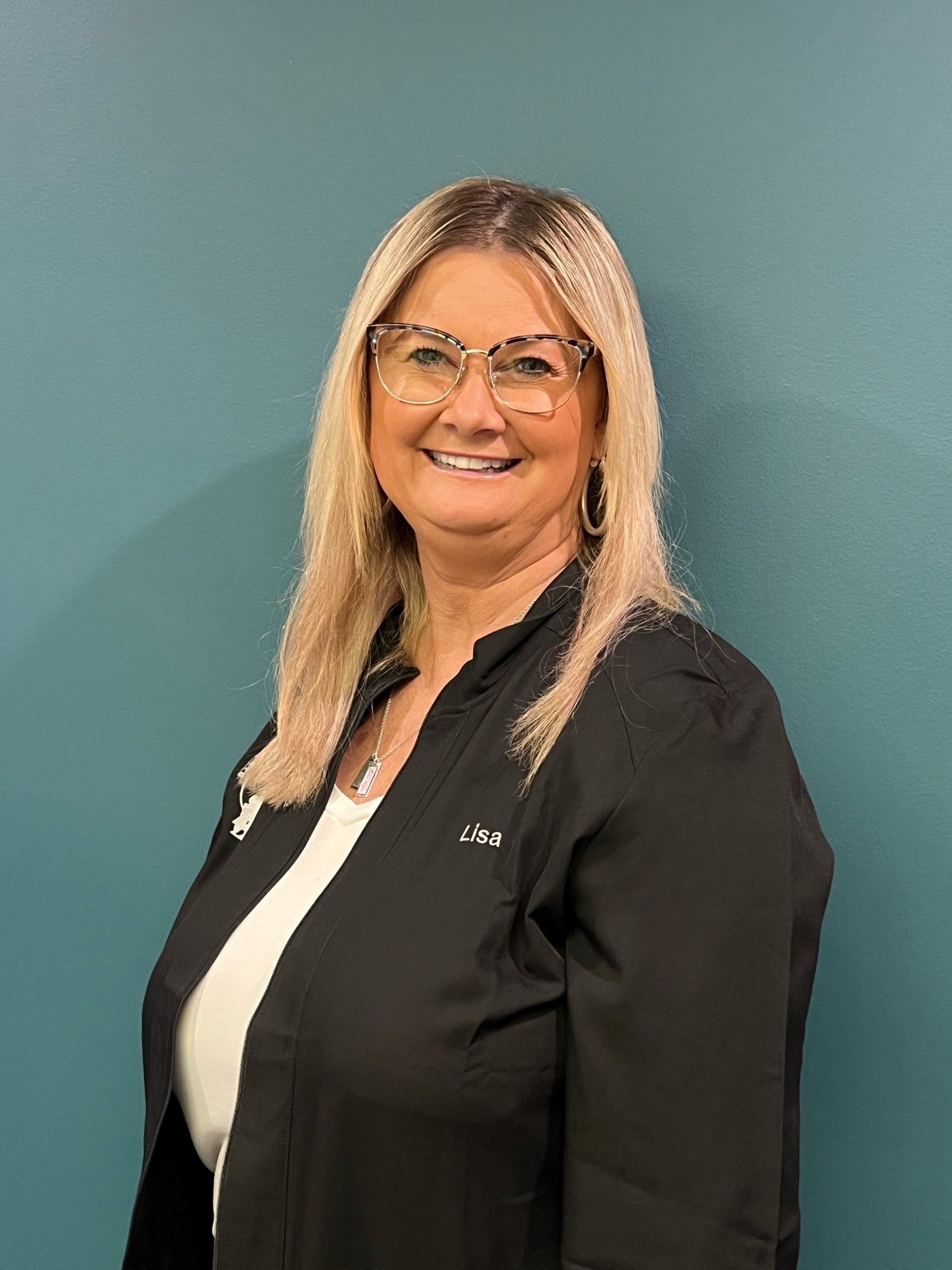 Smiling woman in glasses wearing a black blazer against a teal wall