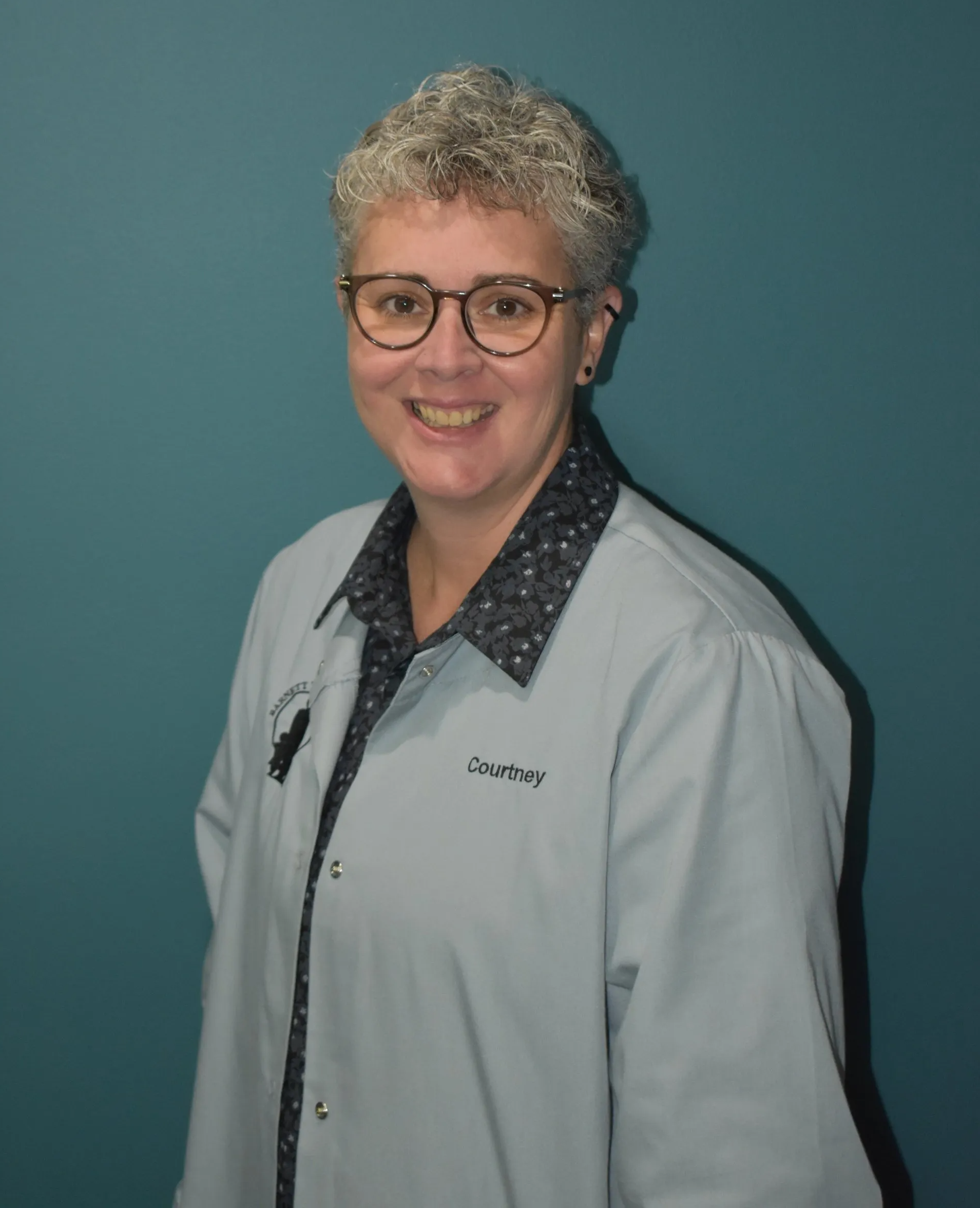 Person in glasses and a light jacket smiling against a teal background.