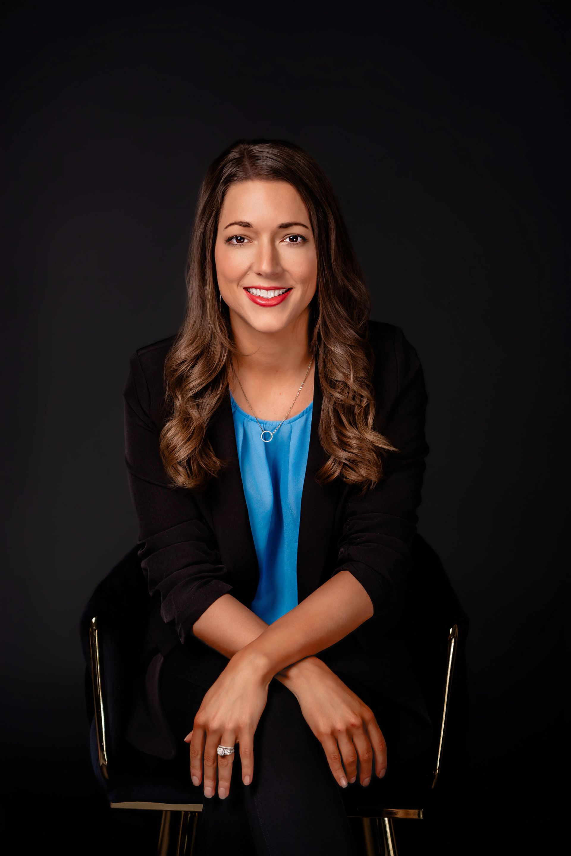 Seated woman in a black blazer and blue blouse, smiling against a dark background
