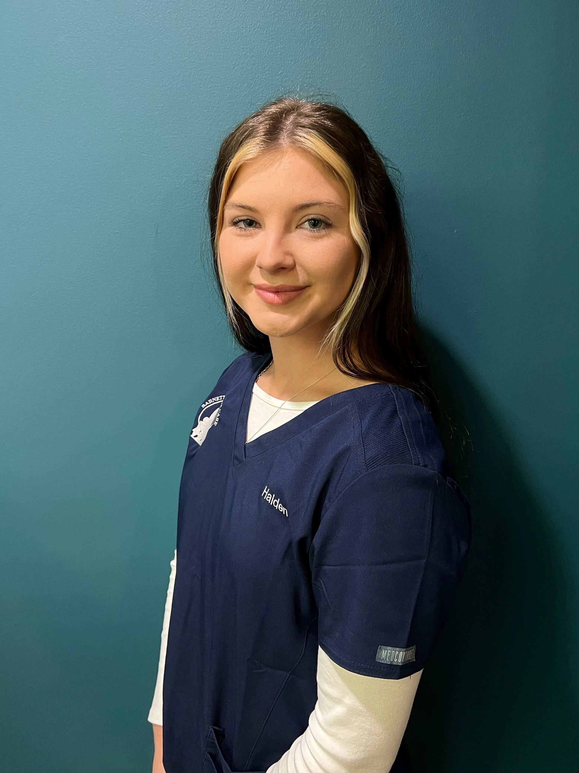 Smiling person in navy scrubs standing against a teal wall