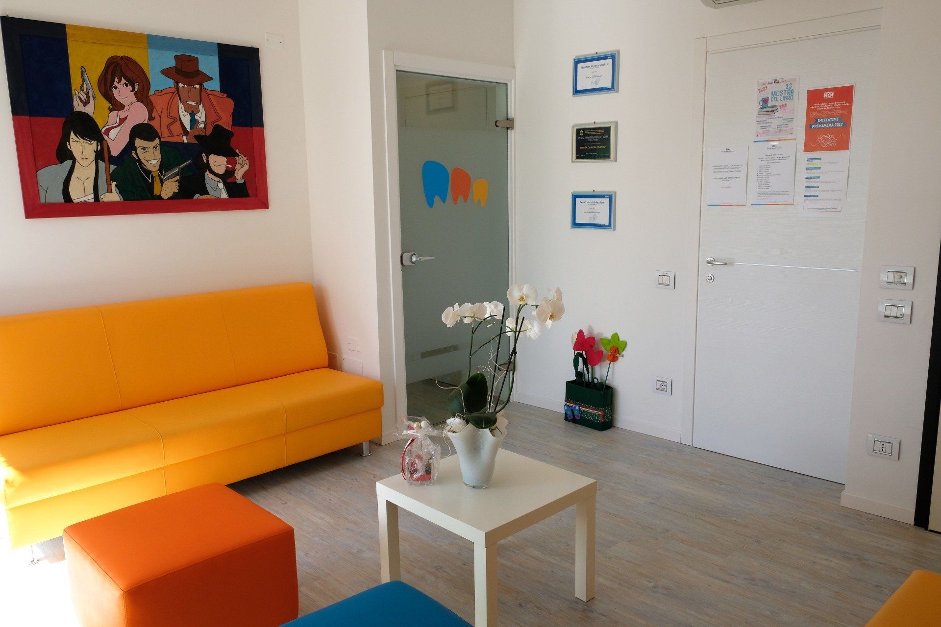 Sala d'attesa con divano arancione, quadri colorati, pareti bianche e una porta a vetri.