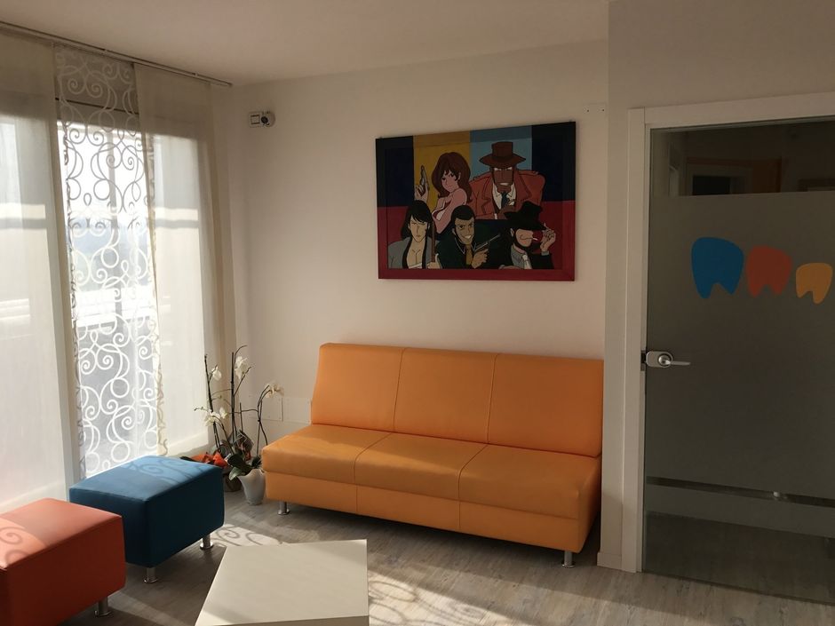 Sala d'attesa con divano arancione, pouf colorati e disegni di cartoni animati; porta in vetro smerigliato con adesivo raffigurante denti.