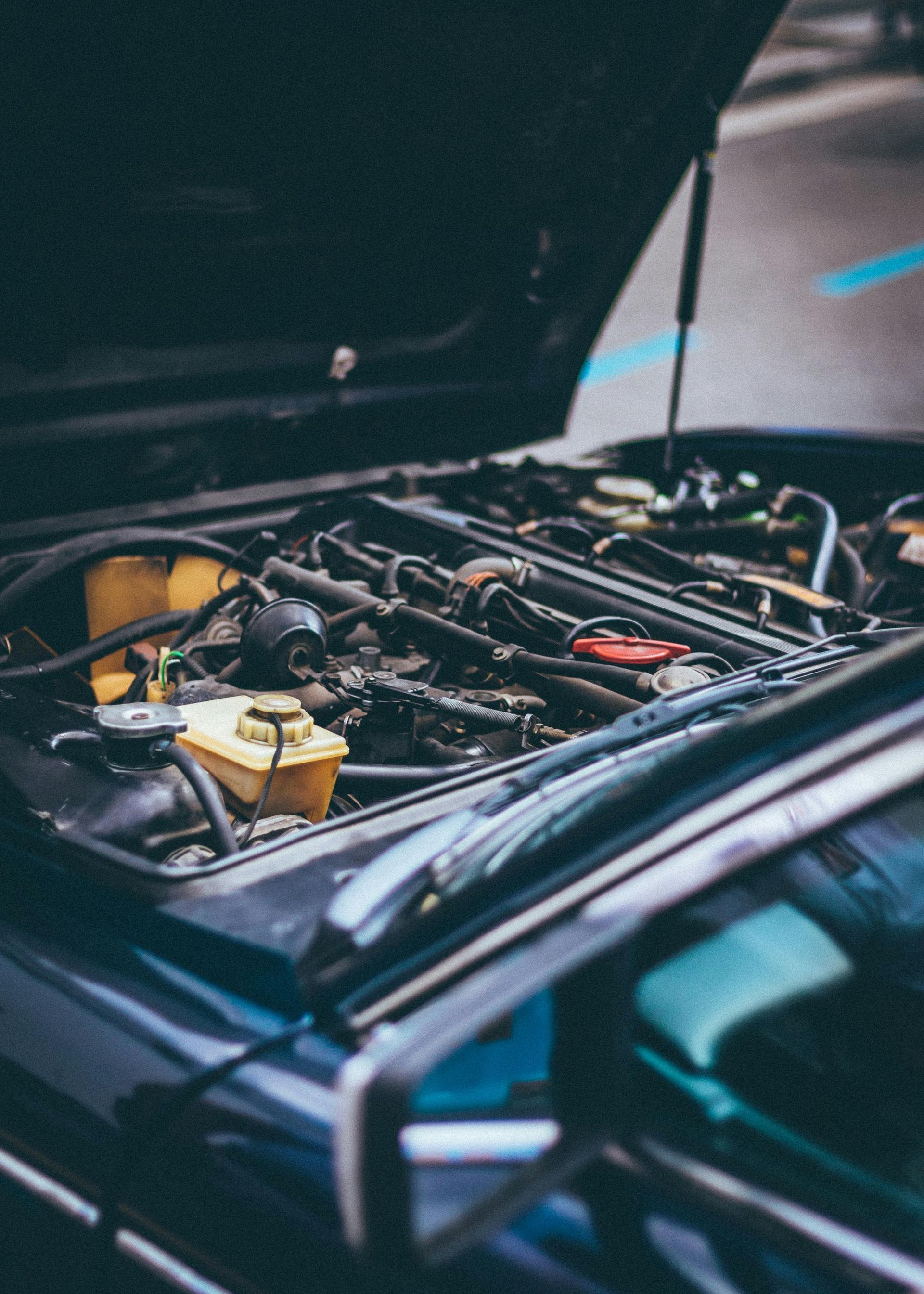 Open car hood revealing a vintage car engine. Dark blue vehicle with various engine components visible.