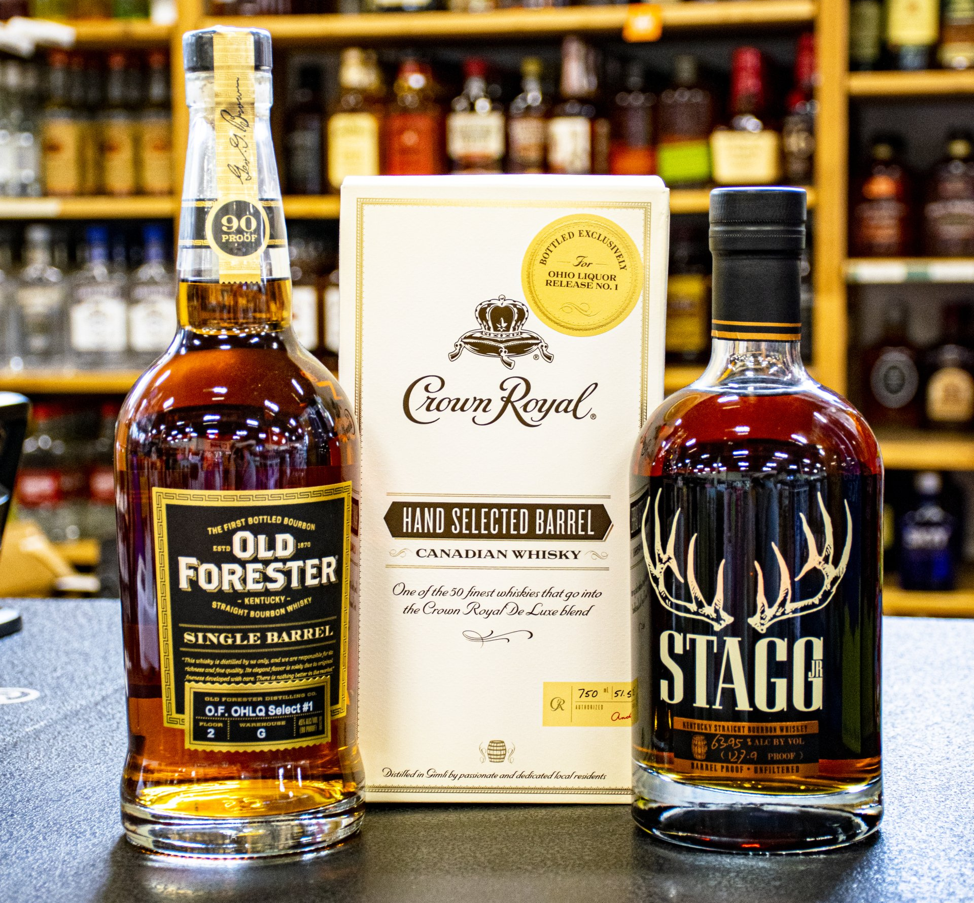 Three whiskey bottles: Old Forester, Crown Royal, and Stagg, displayed in a liquor store setting.