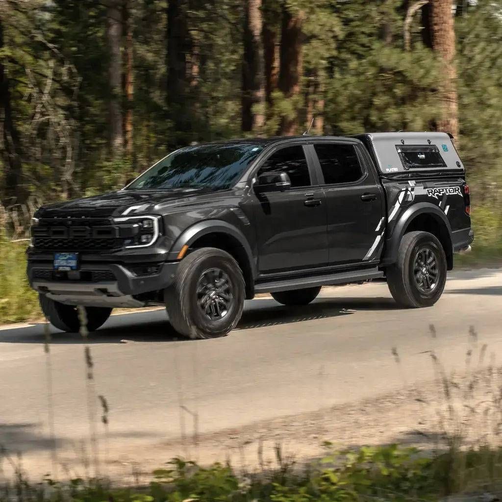 Dark gray Ford Ranger Raptor truck driving on a dirt road in a forest with flated air-topper truck cap.