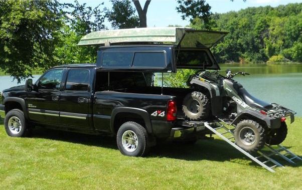 Black pickup truck with Topper EZ lift camper shell, carrying an ATV on a loading ramp near a lake.