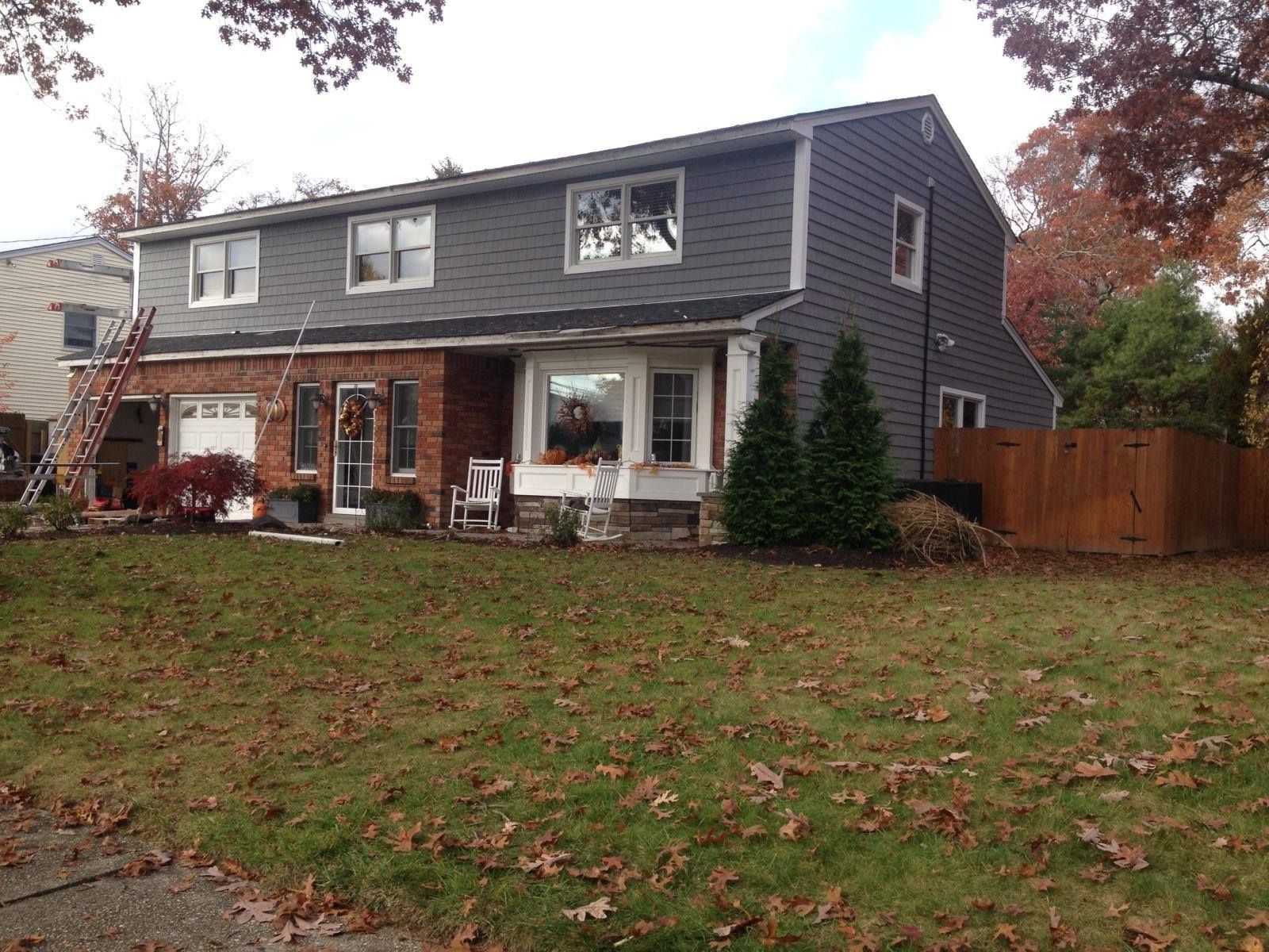 A two-story house with dark gray siding, a brick lower level, a front lawn with fallen leaves, and a person in a window.