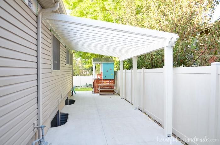 A white patio cover extends from a tan house, shading a concrete walkway alongside a matching white vinyl fence.